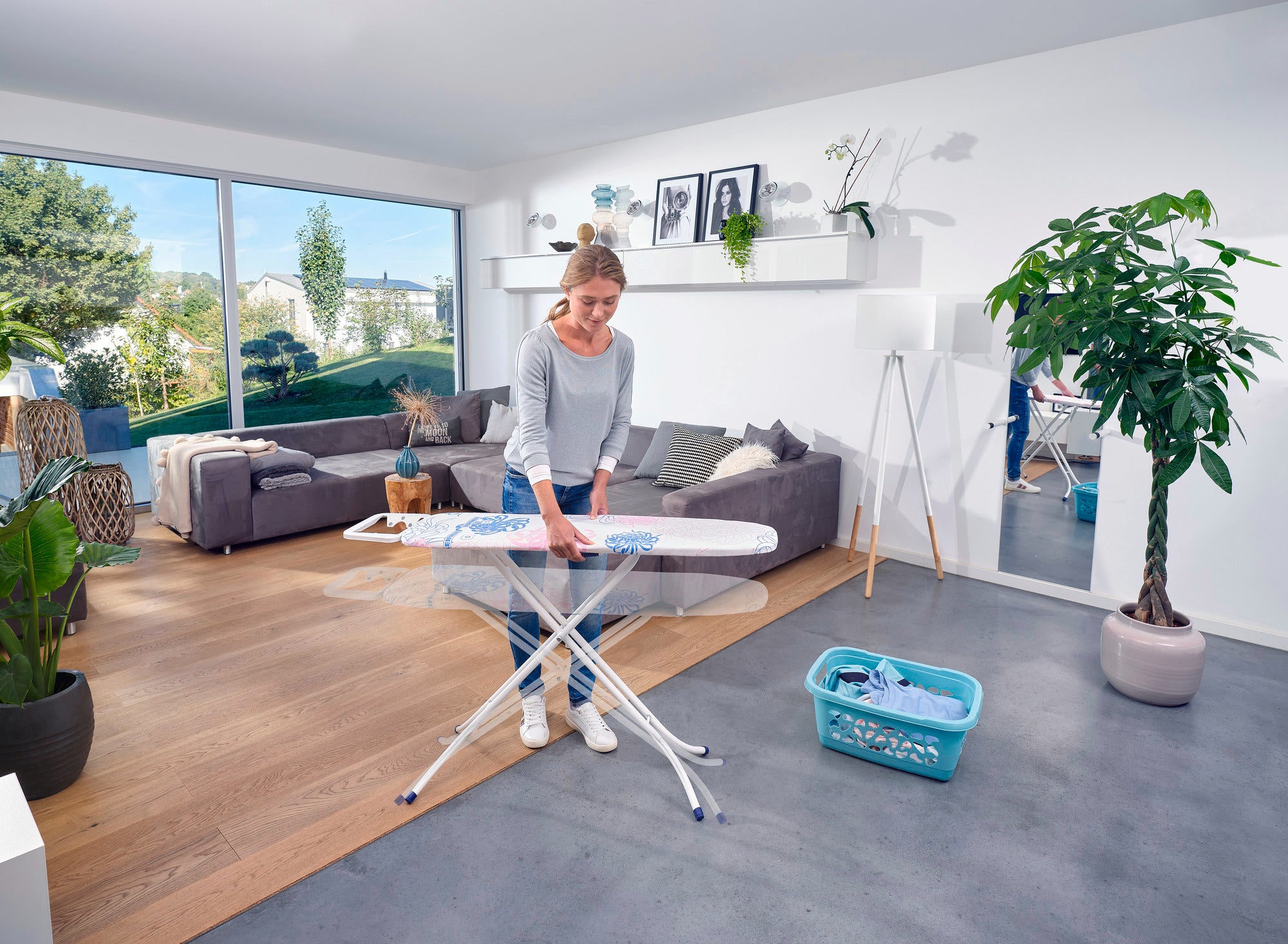 Eine Frau bügelt auf einem Bügeltisch Classic S Basic in einem modernen Wohnzimmer. Der Raum ist hell und stilvoll eingerichtet mit Pflanzen und großen Fenstern.