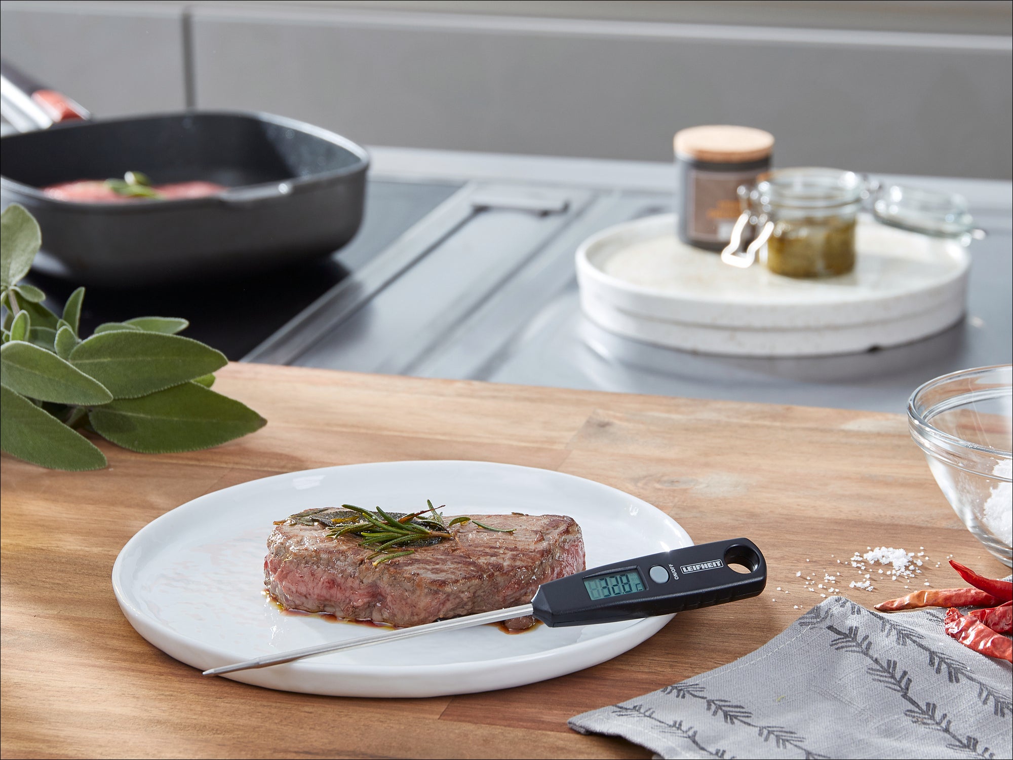 Een digitale keukenthermometer ligt op een bord naast een sappige steak gegarneerd met verse kruiden. Op de achtergrond zijn een pan en kruiden te zien.