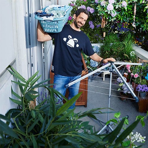 Un homme souriant portant un panier de linge se tient sur un balcon fleuri, pret a etendre les vetements sur un etendoir.