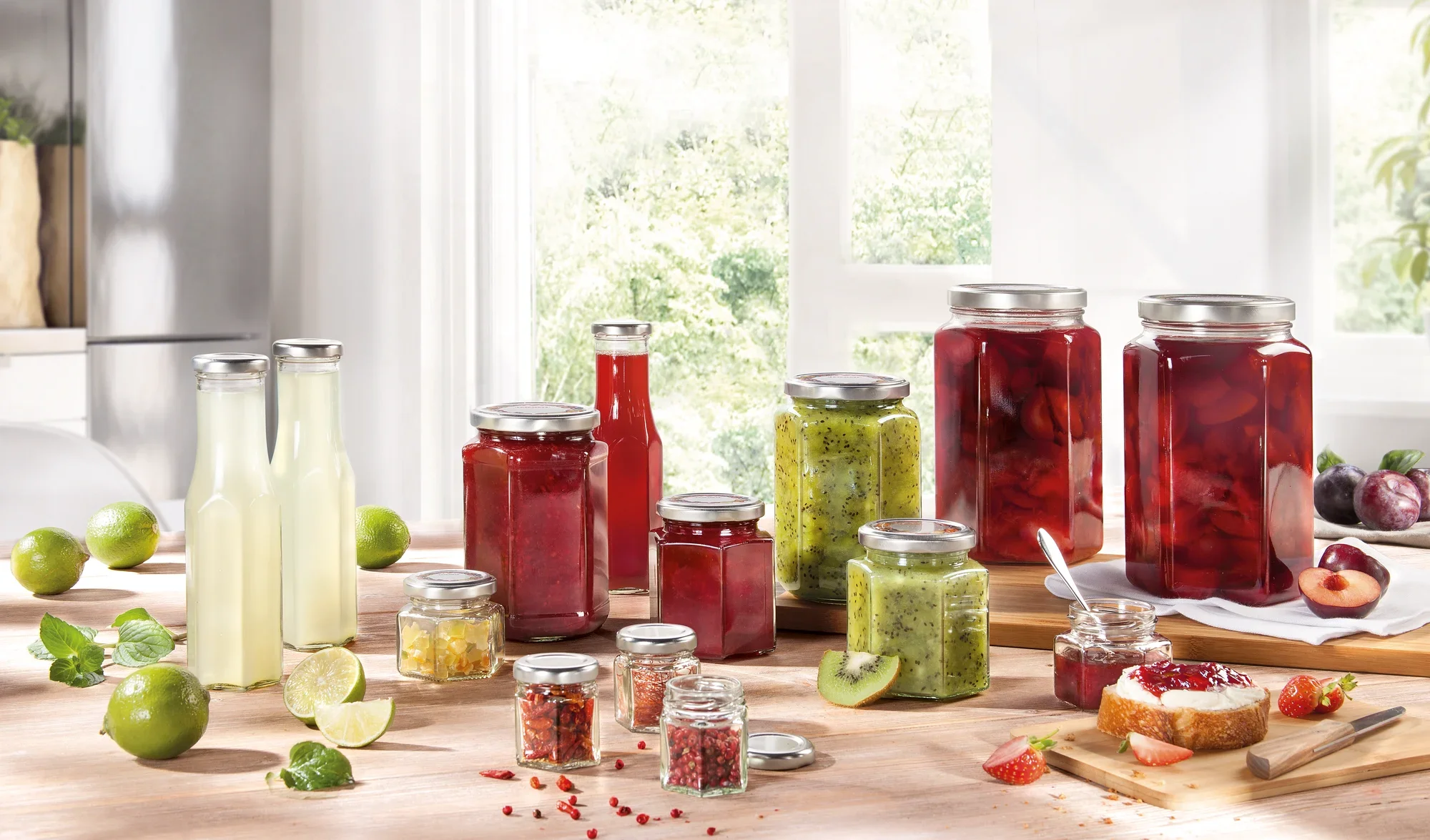 Wybór sześciokątnych słoików wypełnionych różnymi dżemami i sokami stoi na stole. W tle widać okna wychodzące na ogród.