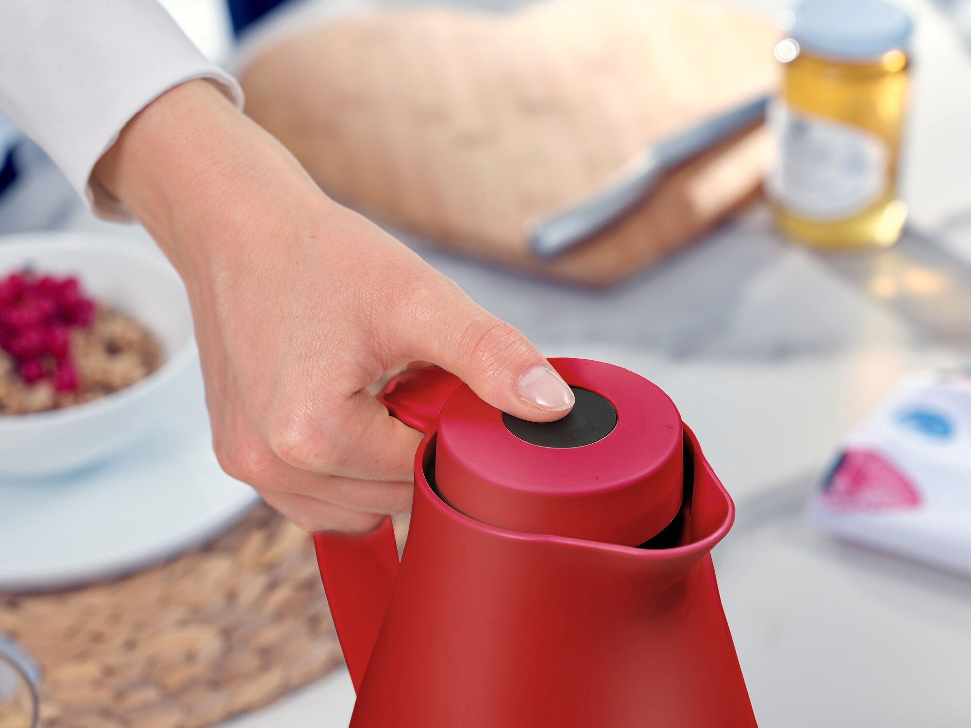 Isolierkanne Harmonic in Rot wird von einer Hand geöffnet. Im Hintergrund sind ein Frühstückstisch mit Müsli und Honig zu sehen. Ideal für stilvolle Heißgetränke.
