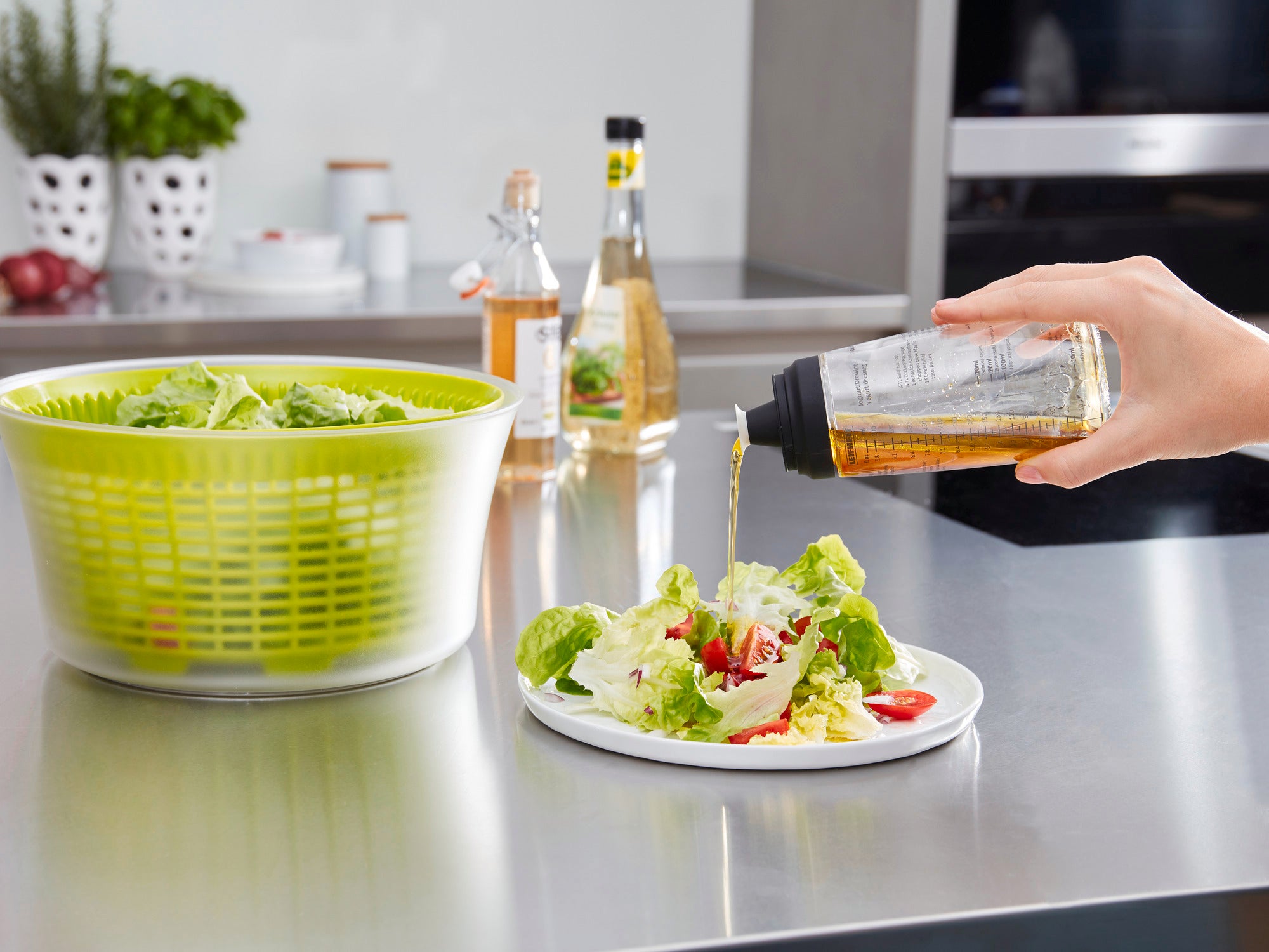 Salat-Dressing-Shaker in Anwendung: Eine Hand gießt Dressing aus einem Shaker über einen frischen Salat auf einem Teller. Im Hintergrund sind eine Salatschleuder und Essigflaschen zu sehen.