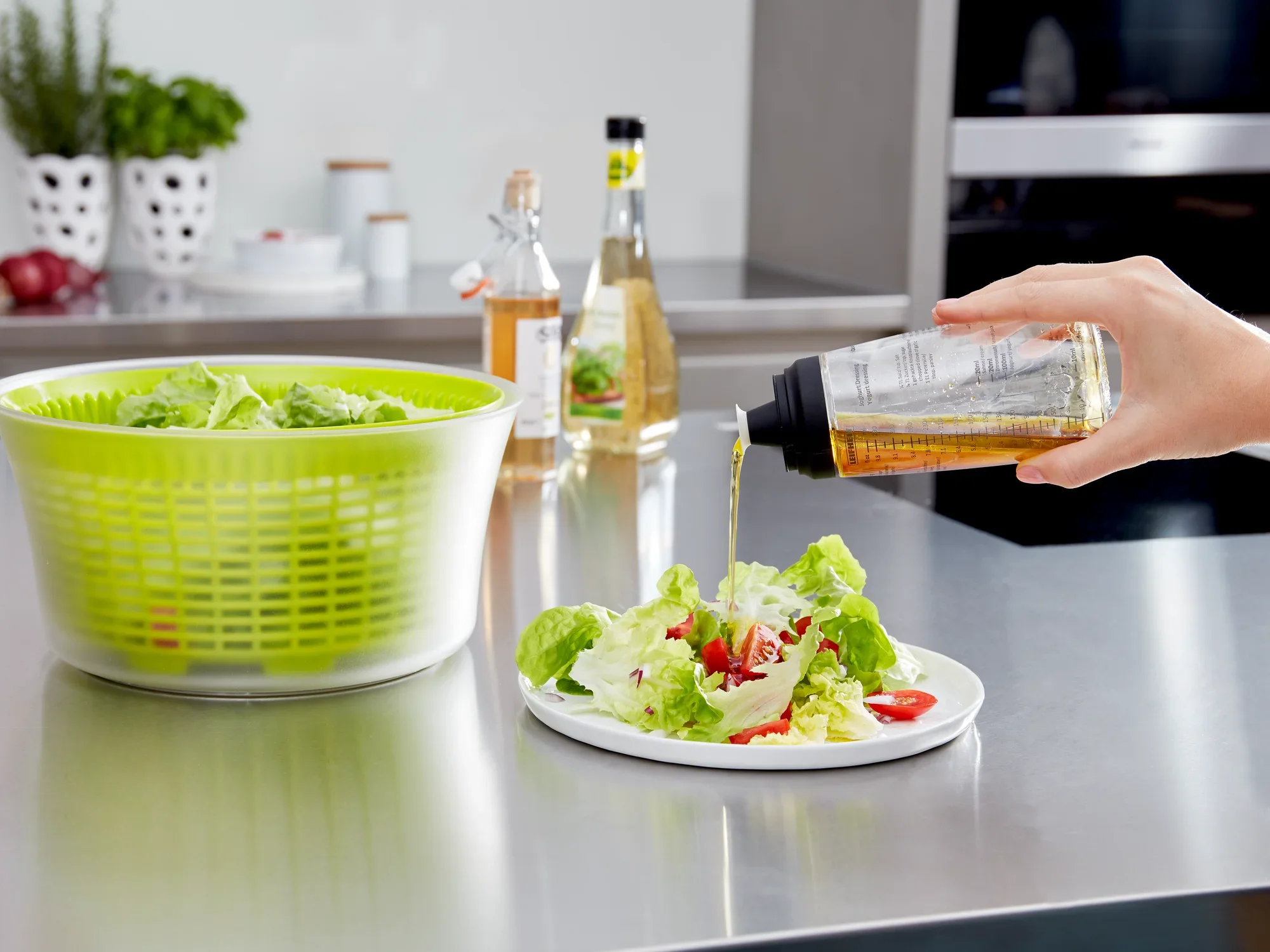 Salade dressing shaker in gebruik: Een hand giet dressing uit een shaker over een verse salade op een bord. Op de achtergrond zie je een slacentrifuge en flessen azijn.