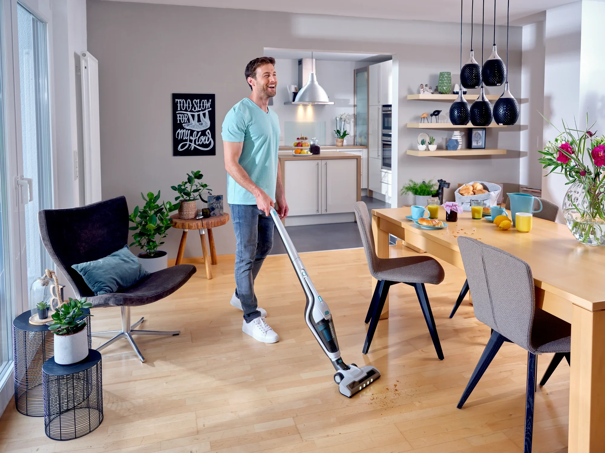 Un homme utilise l'aspirateur sans fil Rotaro PowerVac pour nettoyer le parquet d'un salon moderne et lumineux. La pièce est décorée avec goût, avec des plantes et des objets décoratifs.