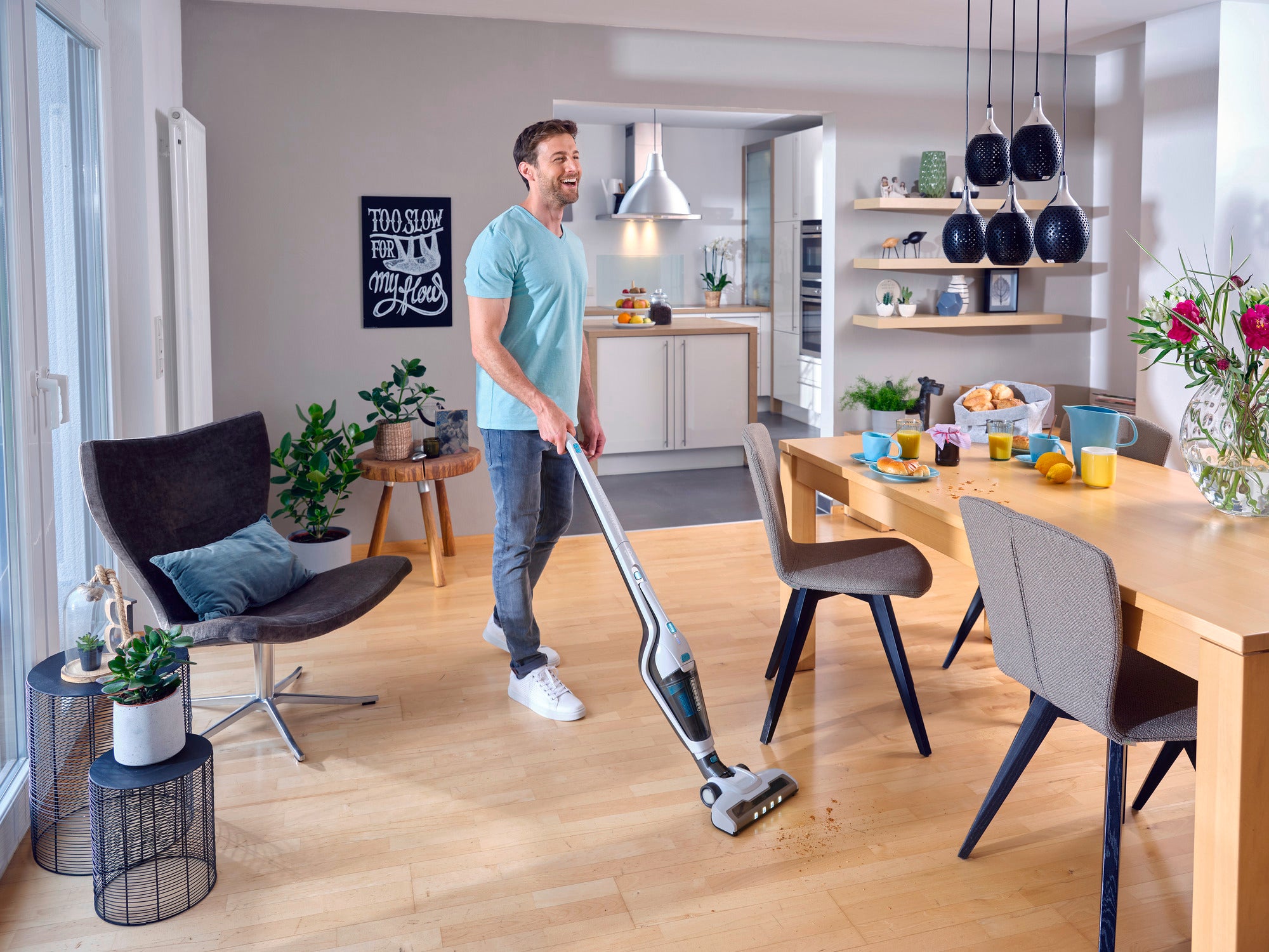 Mężczyzna używa odkurzacza akumulatorowego Rotaro PowerVac do czyszczenia drewnianej podłogi w nowoczesnym, jasnym salonie. Pokój jest stylowo umeblowany roślinami i dekoracjami.