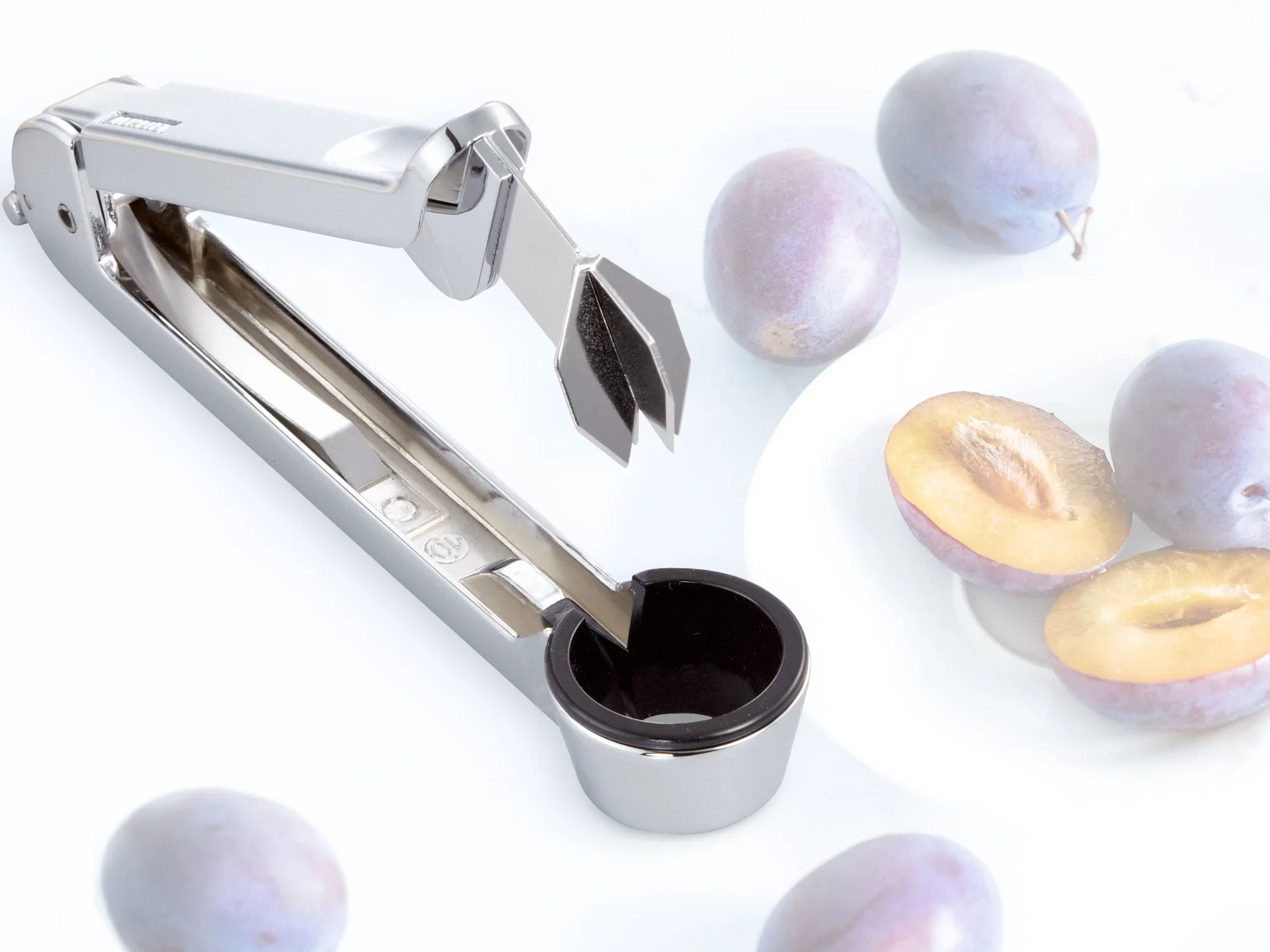 Wydrążacz ze stali nierdzewnej obok świeżych śliwek na białym tle. Narzędzie jest otwarte i gotowe do użycia w celu skutecznego usuwania pestek.