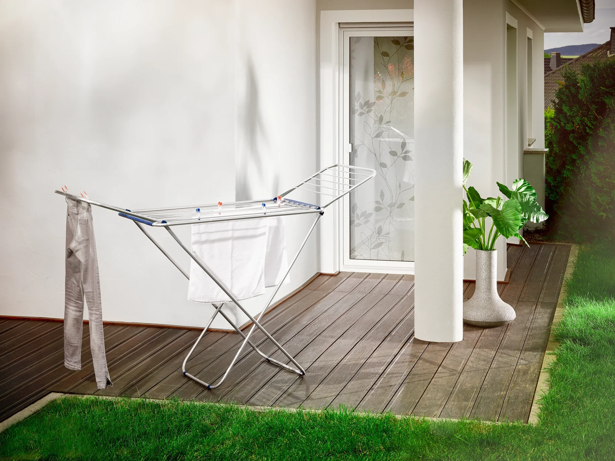 Een Siena 180 vrijstaande droger van aluminium staat op een terras voor een witte huismuur. Op de achtergrond zijn een glazen deur en een plant in een witte pot te zien.