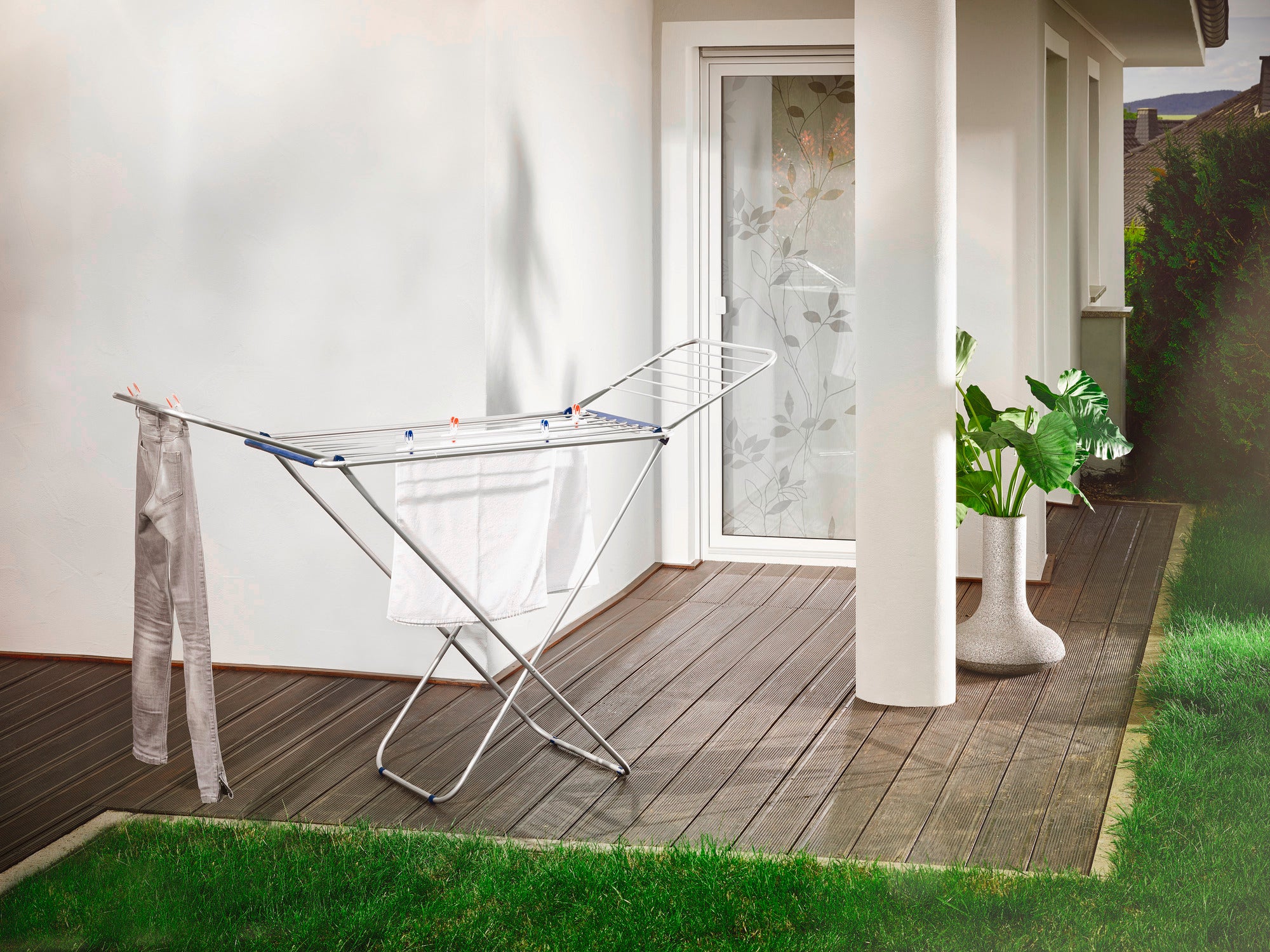 Een Siena 180 vrijstaande droger van aluminium staat op een terras voor een witte huismuur. Op de achtergrond zijn een glazen deur en een plant in een witte pot te zien.