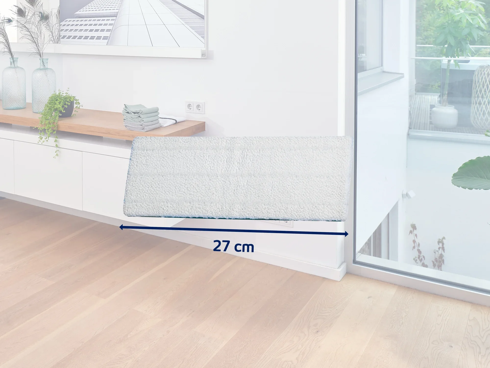 Ein weißer, rechteckiger Wischmopp mit einer Breite von 27 cm liegt auf einem hellen Holzboden in einem modernen, lichtdurchfluteten Raum. Im Hintergrund sind ein Sideboard und Pflanzen zu sehen.