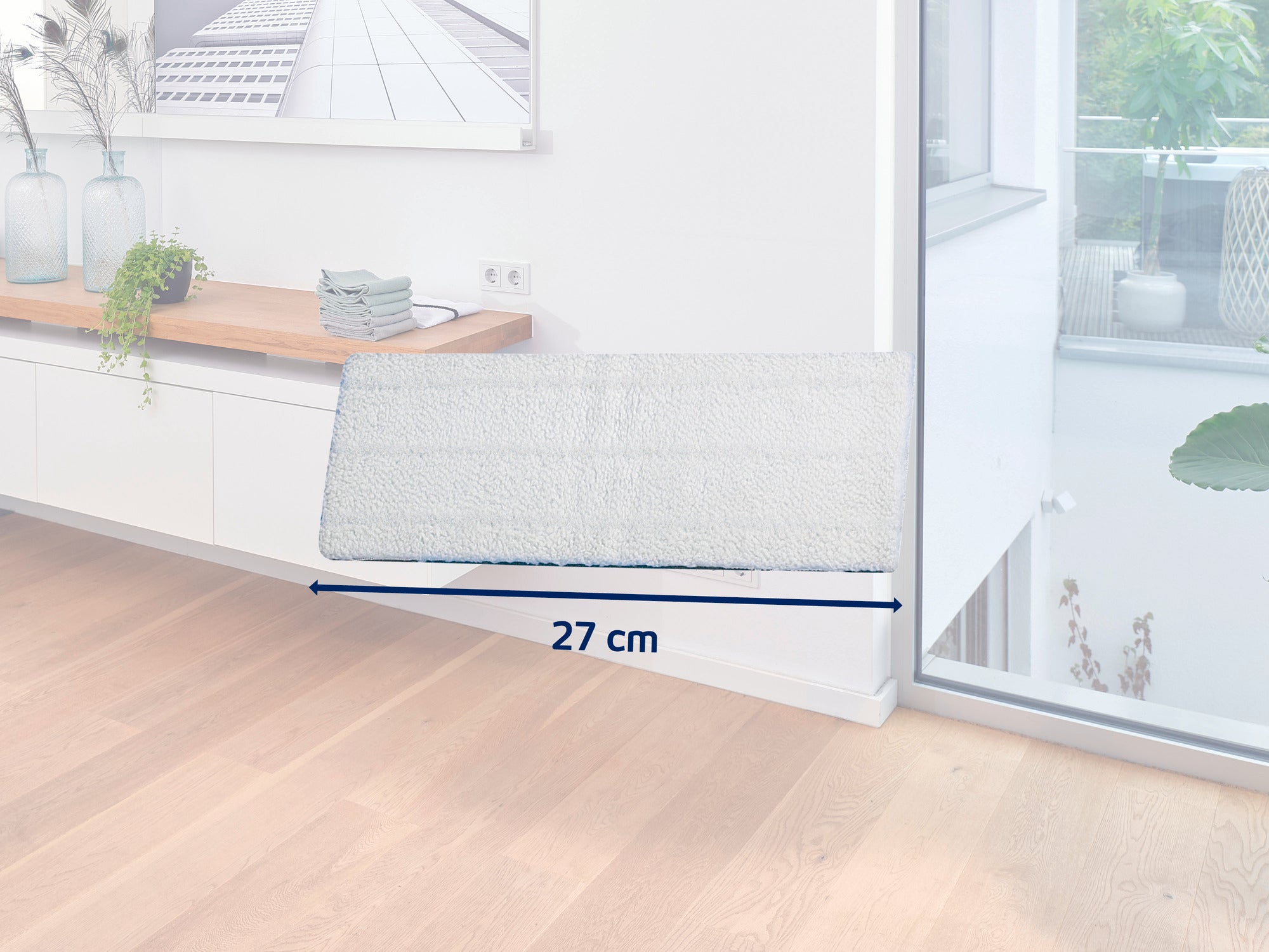 Biały, prostokątny mop o szerokości 27 cm leży na jasnej drewnianej podłodze w nowoczesnym, jasnym pokoju. W tle widać kredens i rośliny.