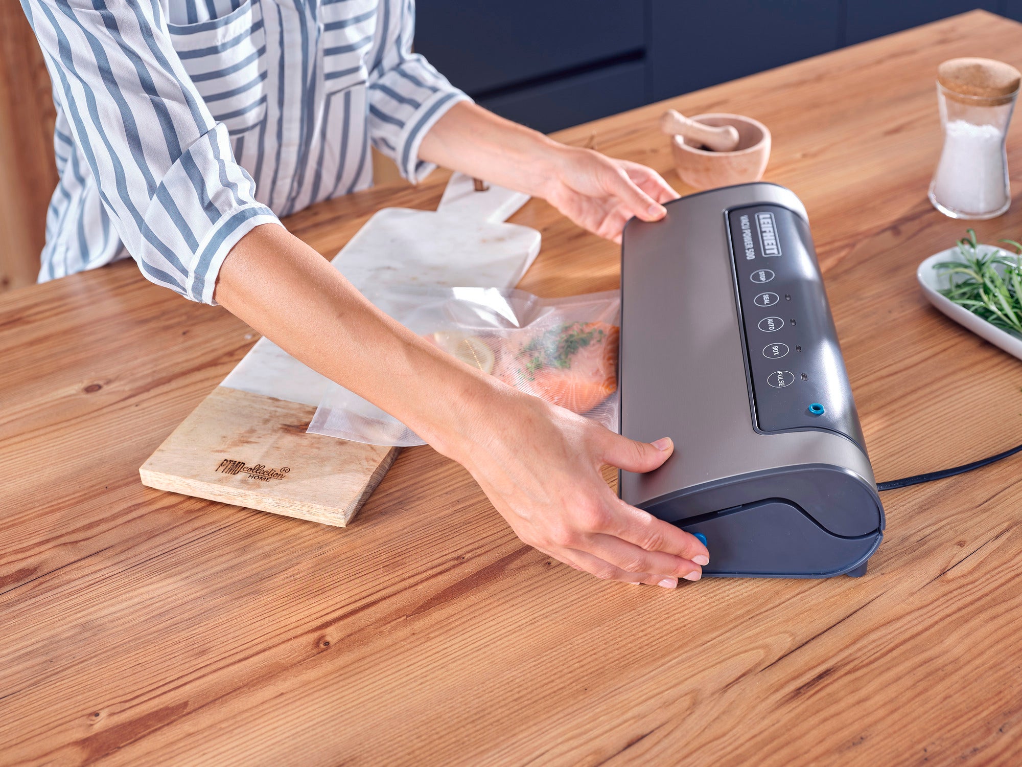 Une personne utilise l'appareil de mise sous vide Vacu Power 500 pour sceller des aliments dans un sachet posé sur une table en bois. L'appareil est compact et facile à utiliser.