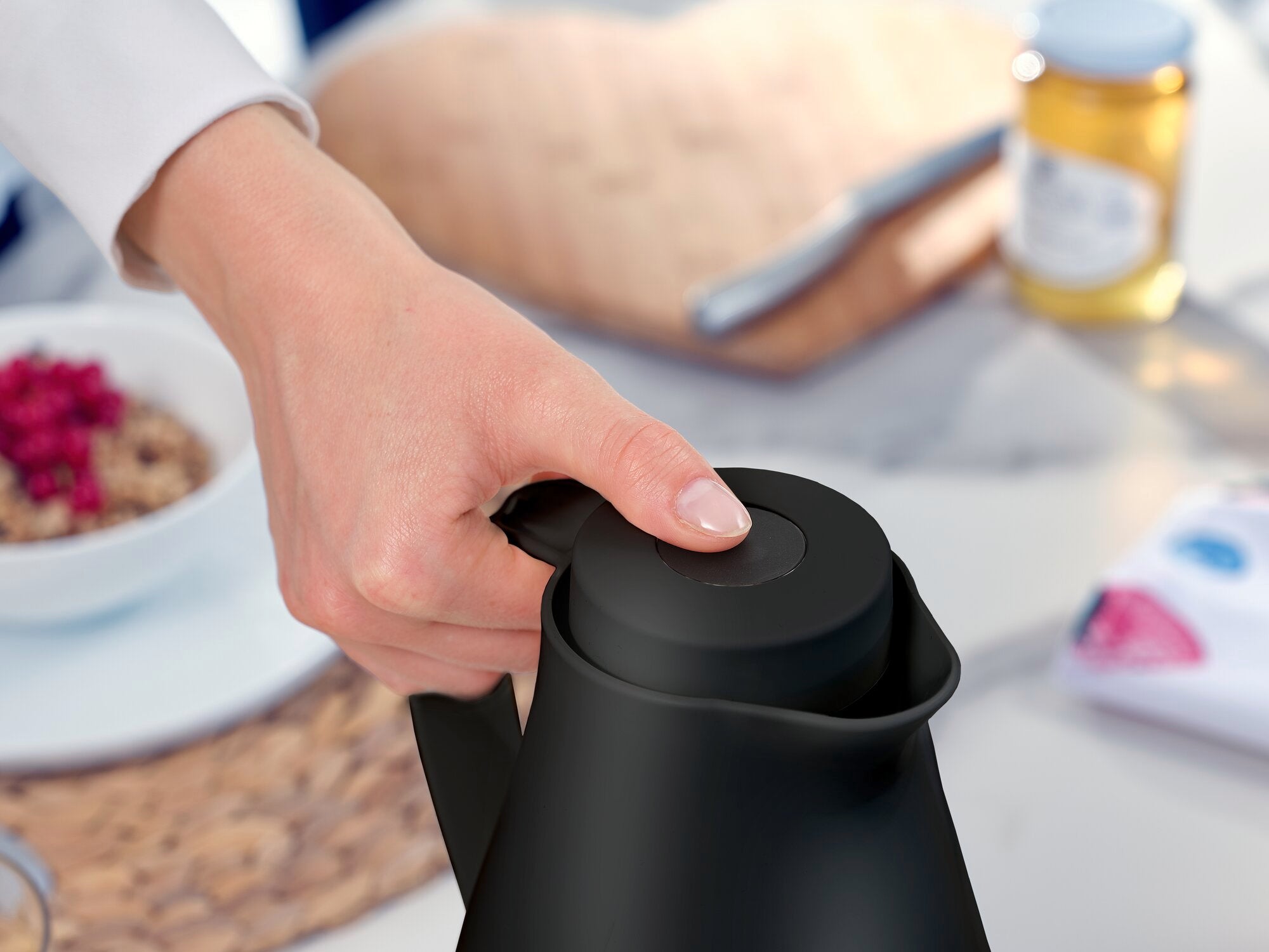 Eén hand houdt het deksel van de zwarte "Harmonic" vacuümfles vast. Op de achtergrond is een gedekte tafel met ontbijtgerei en een pot honing te zien.