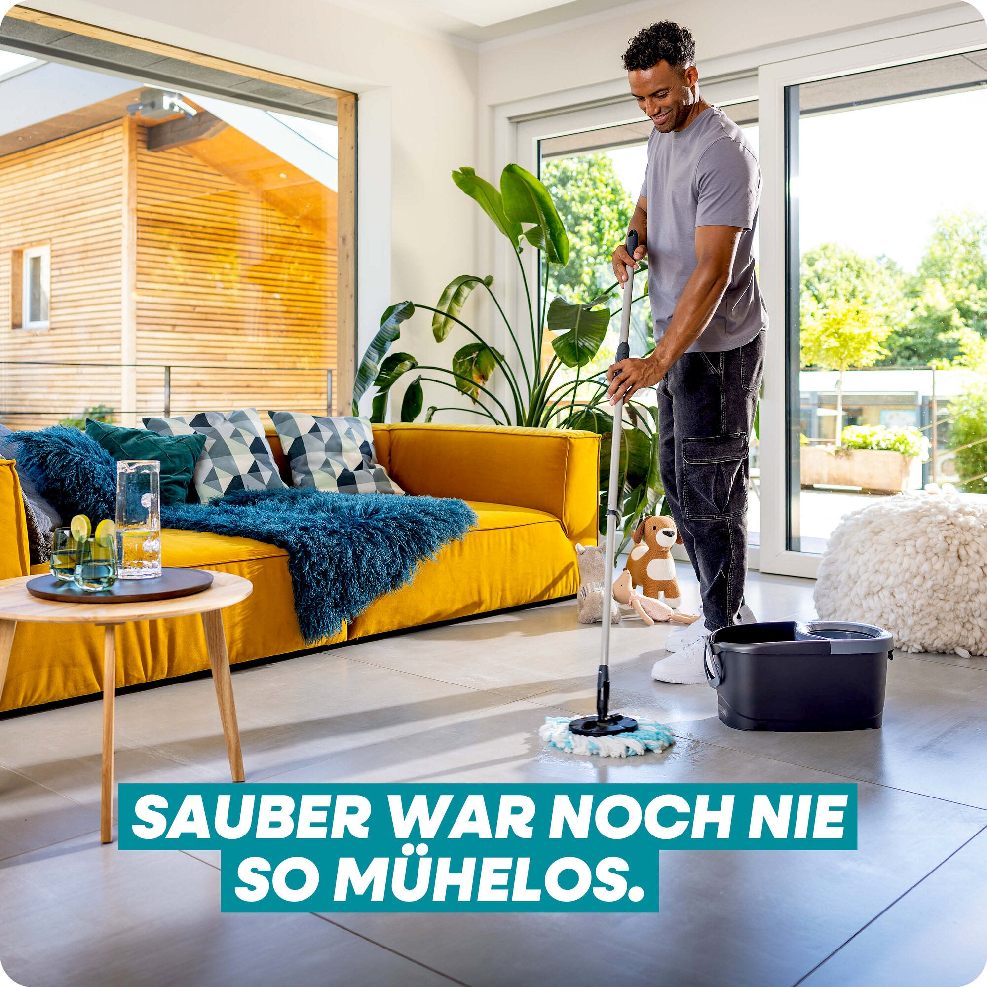 Leifheit Set CLEAN TWIST Disc Mop Black im Einsatz: Ein Mann reinigt mühelos den hellen Wohnraumboden mit Rundmopp und Eimer neben gelbem Sofa und großen Fenstern.