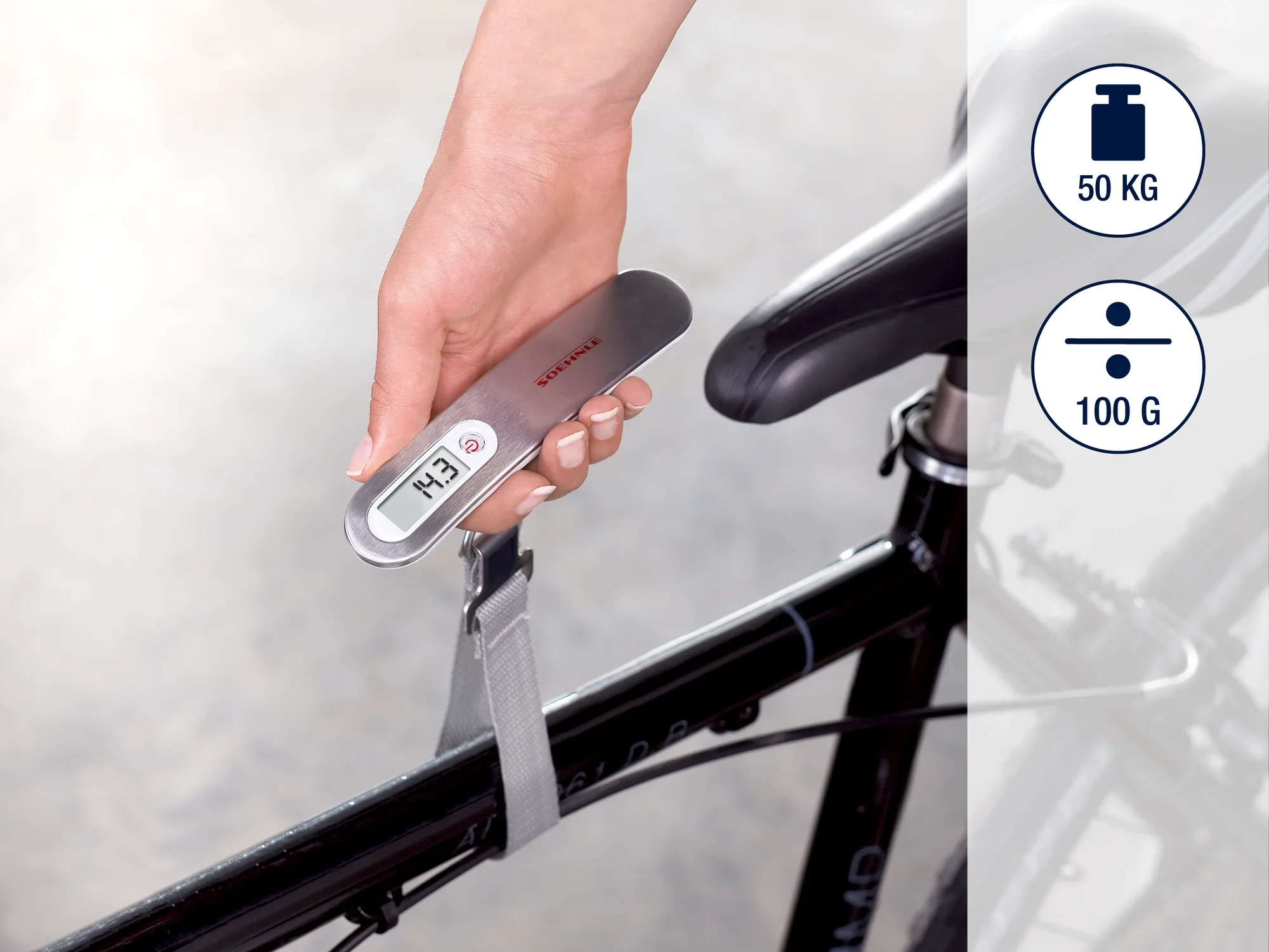 Jedna ręka trzyma cyfrową wagę bagażową, która jest przymocowana do ramy roweru. Waga wyświetla wagę w kilogramach i ma maksymalny udźwig 50 kg.