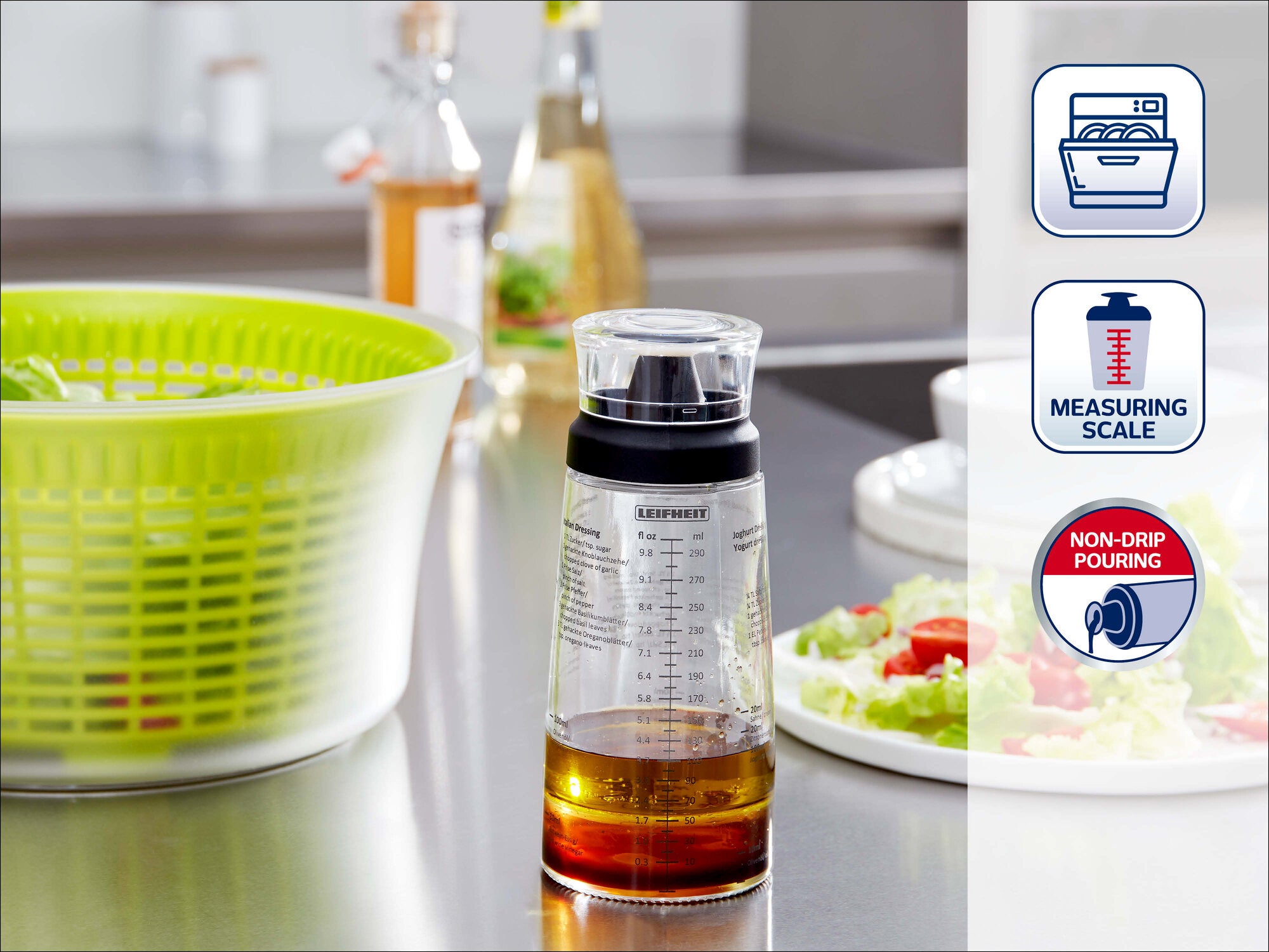 Leifheit Dressing Shaker mit integrierter Messskala und tropffreiem Ausgießer, gefüllt mit Dressing auf der Küchenarbeitsplatte; daneben Salatschüssel, Teller und Ölflaschen.