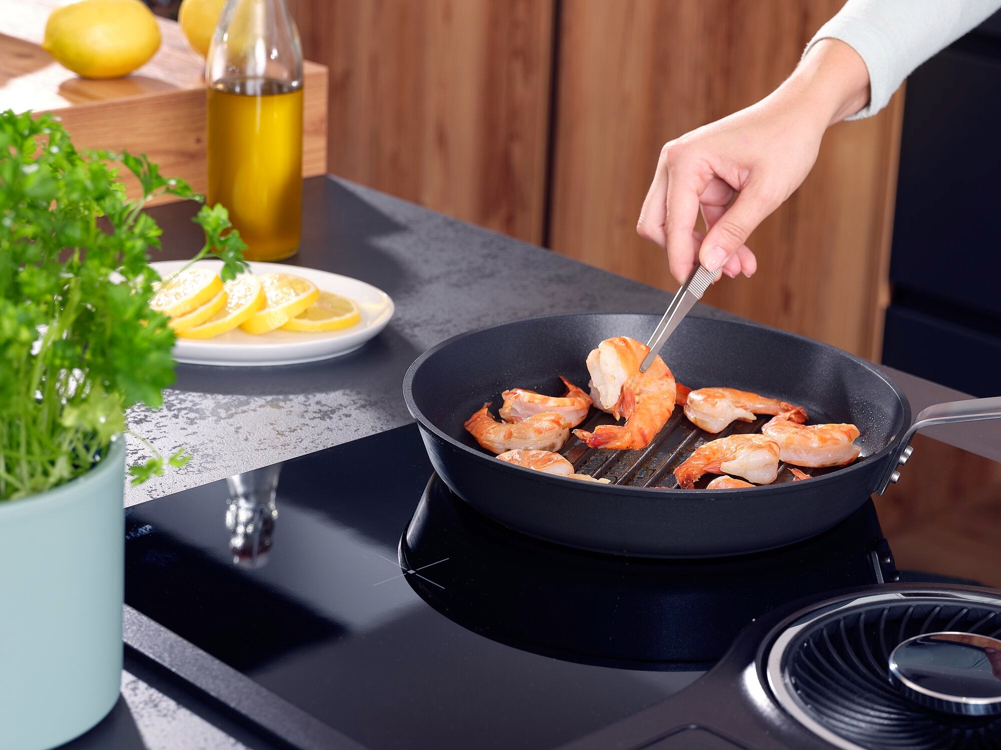 Pinzetta da cucina 20 cm: una mano tiene una pinzetta in acciaio inossidabile e gira i gamberetti in una padella sul fornello. Sullo sfondo si vedono limoni ed erbe aromatiche.