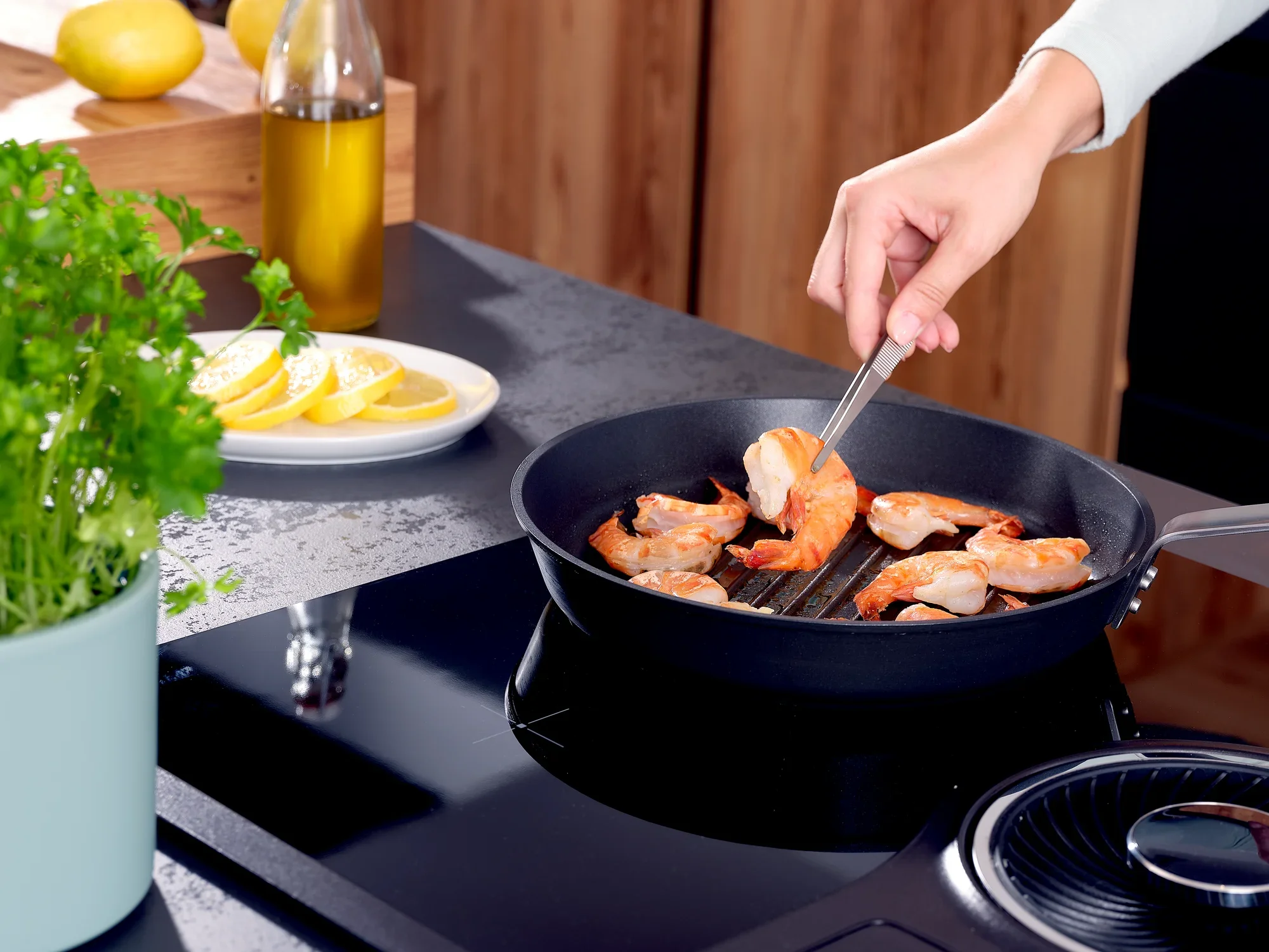 Keukenpincet 20 cm: Een hand houdt een roestvrijstalen pincet vast en draait garnalen in een pan op een kookplaat. Op de achtergrond zijn citroenen en kruiden te zien.