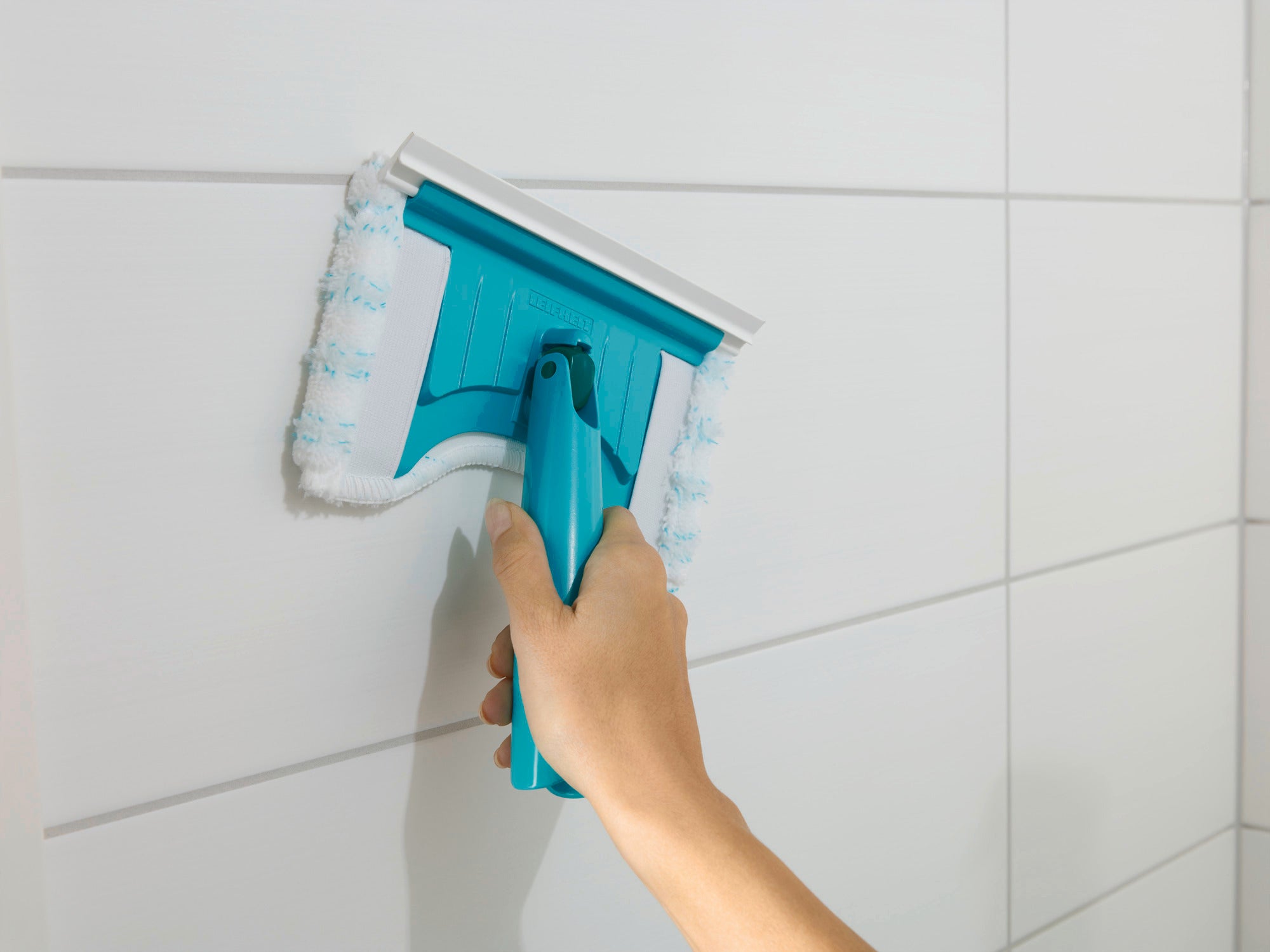A Flexipad tile and tub squeegee with telescopic handle is used to clean white tiles. The squeegee has a blue handle and a white cleaning pad.