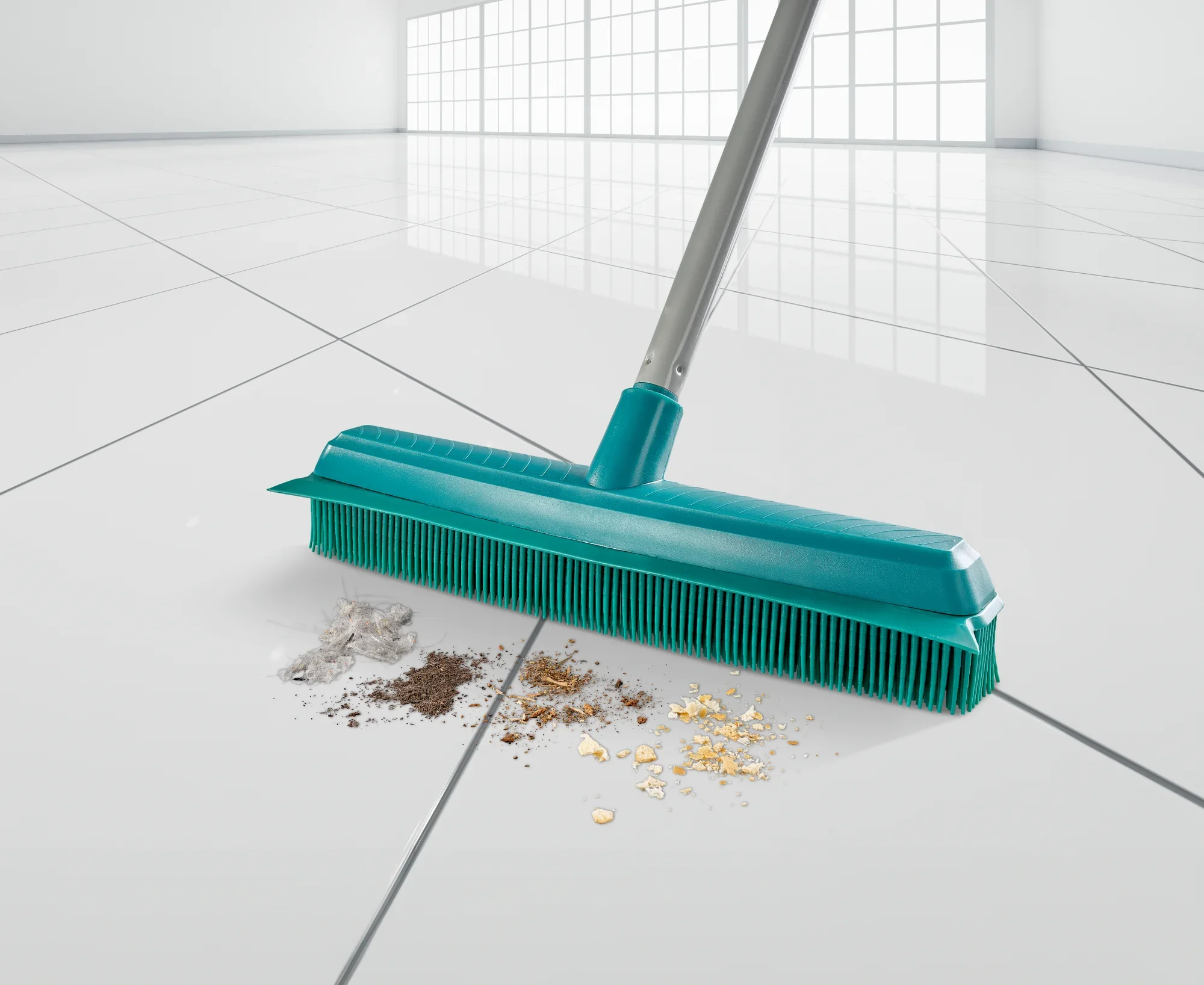 Una scopa Suprabesen turchese pulisce efficacemente un pavimento di piastrelle bianche lucide. La scopa rimuove sporco, polvere e piccole particelle per garantire una pulizia impeccabile.