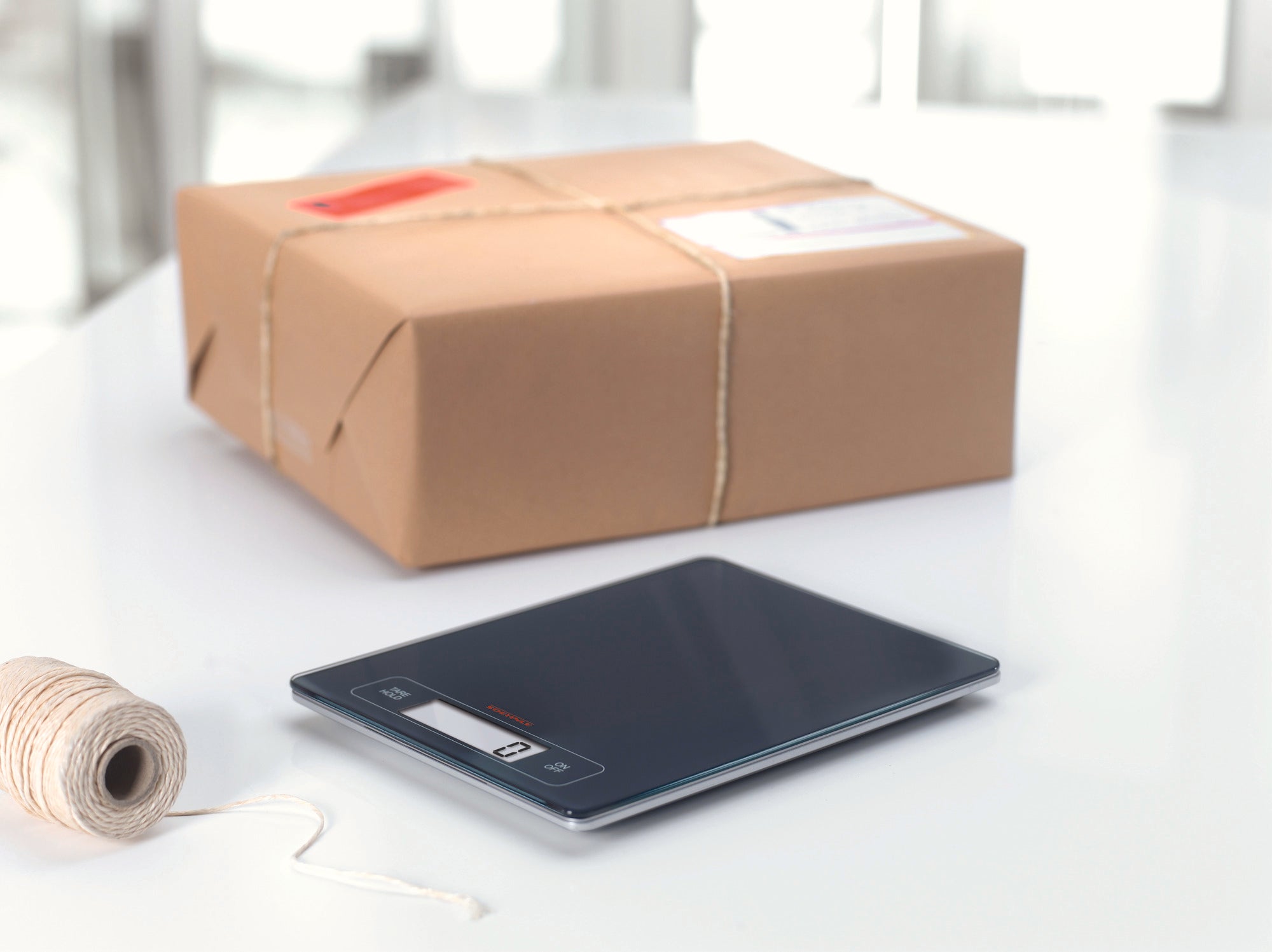 Eine digitale Küchenwaage neben einem verpackten Karton auf einem weißen Tisch. Die Waage hat ein modernes Design mit einem klaren Display, ideal für präzises Wiegen.