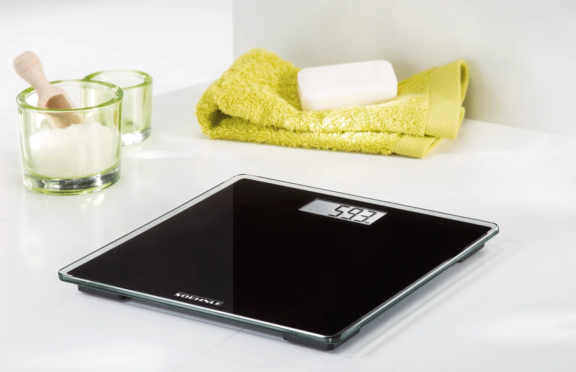 Personenwaage digital Style Sense Compact 100 in elegantem Schwarz auf einem weißen Tisch. Im Hintergrund sind gelbe Handtücher und Seife zu sehen. Ideal für modernes Badezimmerdesign.