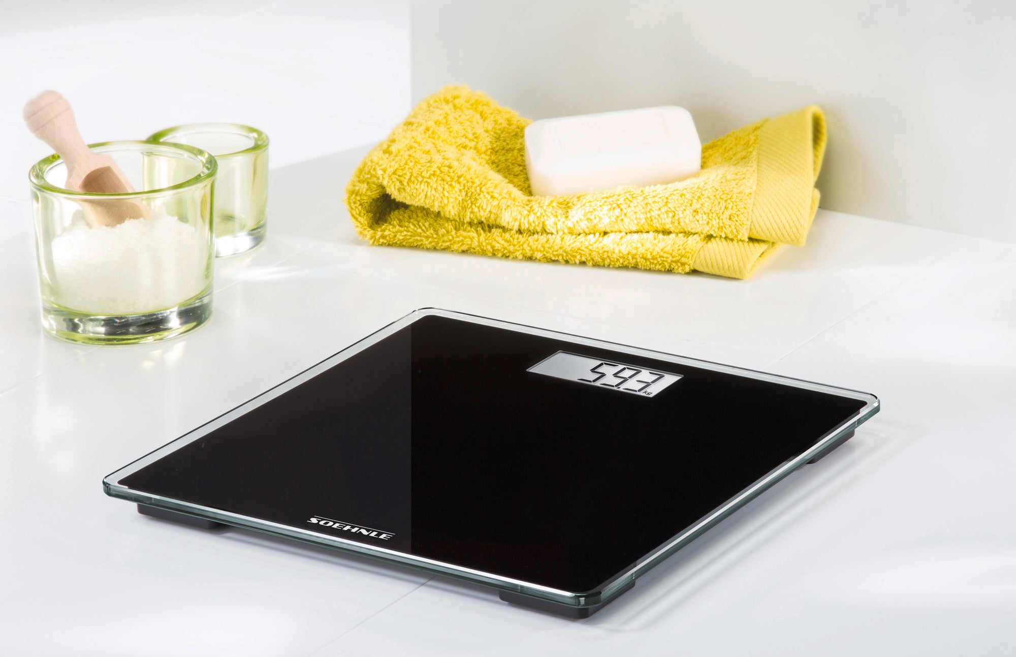 Personenwaage digital Style Sense Compact 100 in elegantem Schwarz auf einem weißen Tisch. Im Hintergrund sind gelbe Handtücher und Seife zu sehen. Ideal für modernes Badezimmerdesign.