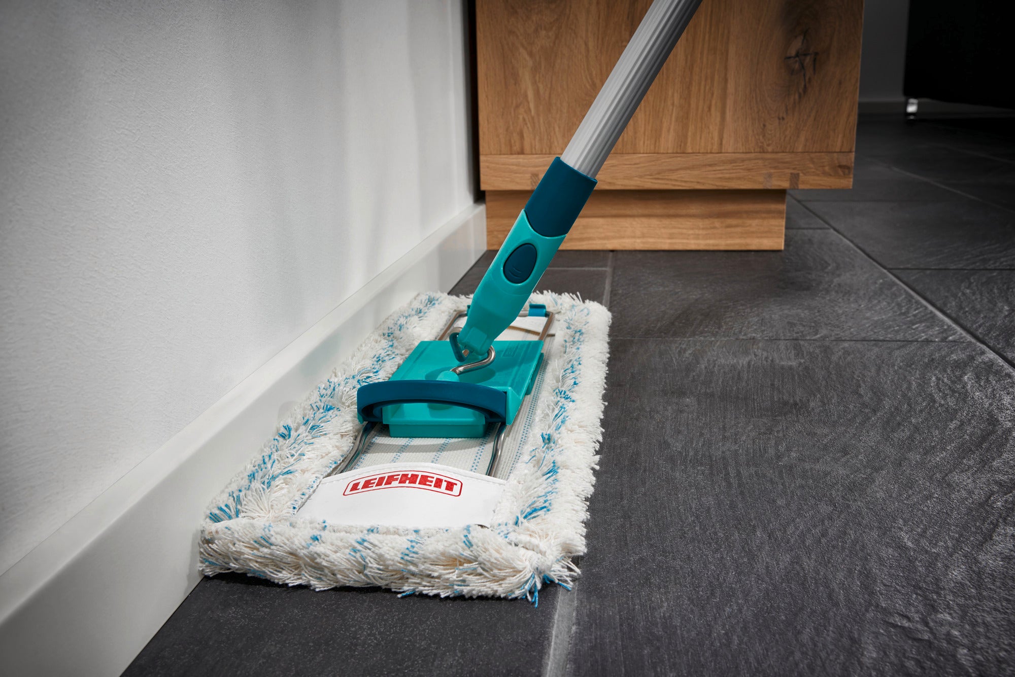 Bodenwischer Profi Cotton Plus in Aktion auf einem dunklen Fliesenboden. Der Wischer hat einen türkisfarbenen Griff und ein weißes, flauschiges Wischtuch. Ideal für gründliche Reinigung.