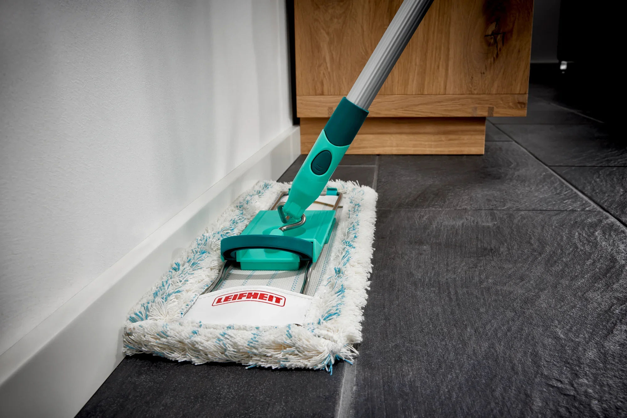 Balai Profi Cotton Plus en action sur un sol carrelé foncé. Le balai a un manche turquoise et une serpillière blanche et moelleuse. Idéal pour un nettoyage en profondeur.
