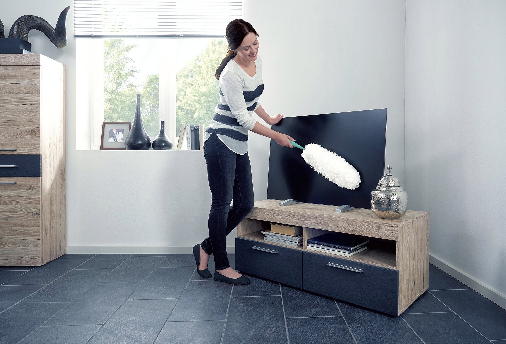 Eine Frau benutzt den Staubwedel, um einen Flachbildfernseher in einem modernen Wohnzimmer zu reinigen. Der Staubwedel ist weiß und effektiv für glatte Oberflächen.