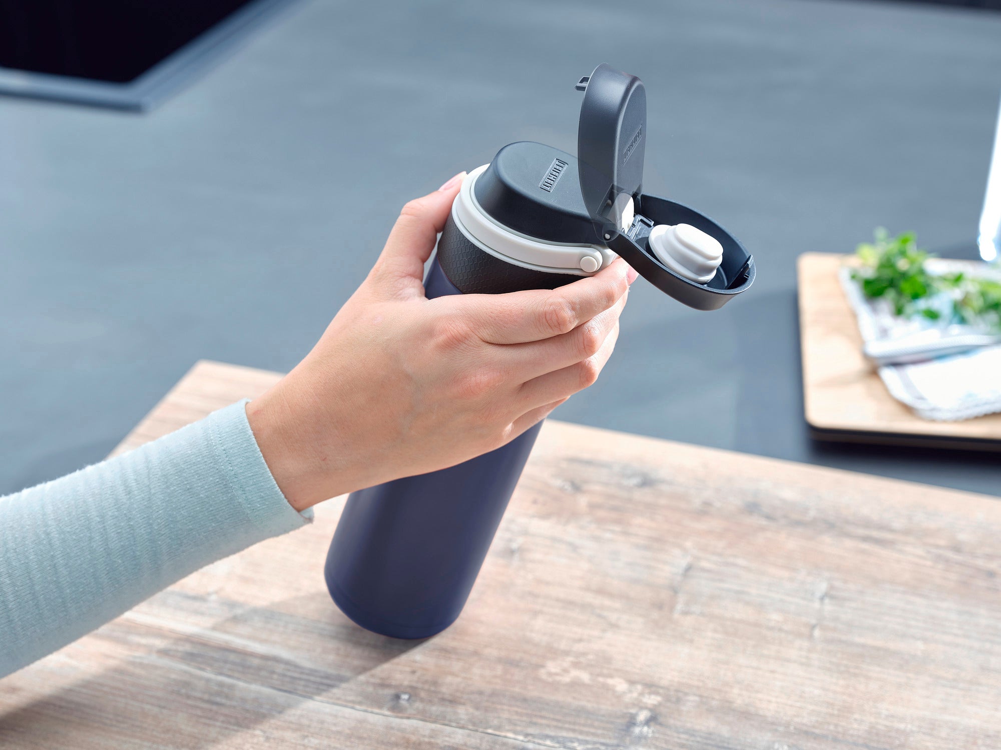 Isolierbecher Flip 600ml in Dunkelblau wird auf einem Tisch gehalten. Der Deckel ist geöffnet, um das praktische Design und die einfache Handhabung zu zeigen. Ideal für unterwegs.