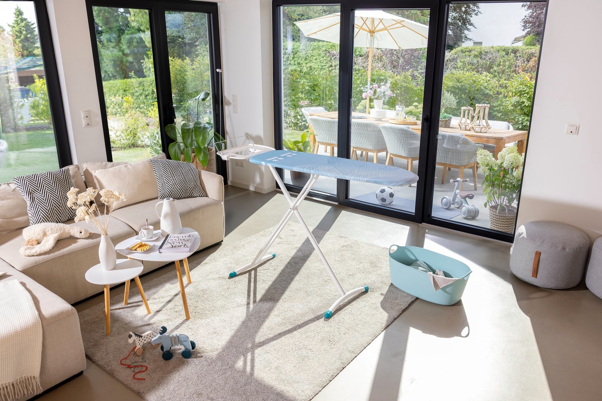Bügelbrett Leifheit Air Board Express M Solid in einem modernen Wohnzimmer mit großen Fenstern. Es steht auf einem hellen Teppich neben einem Sofa und einem Korb.