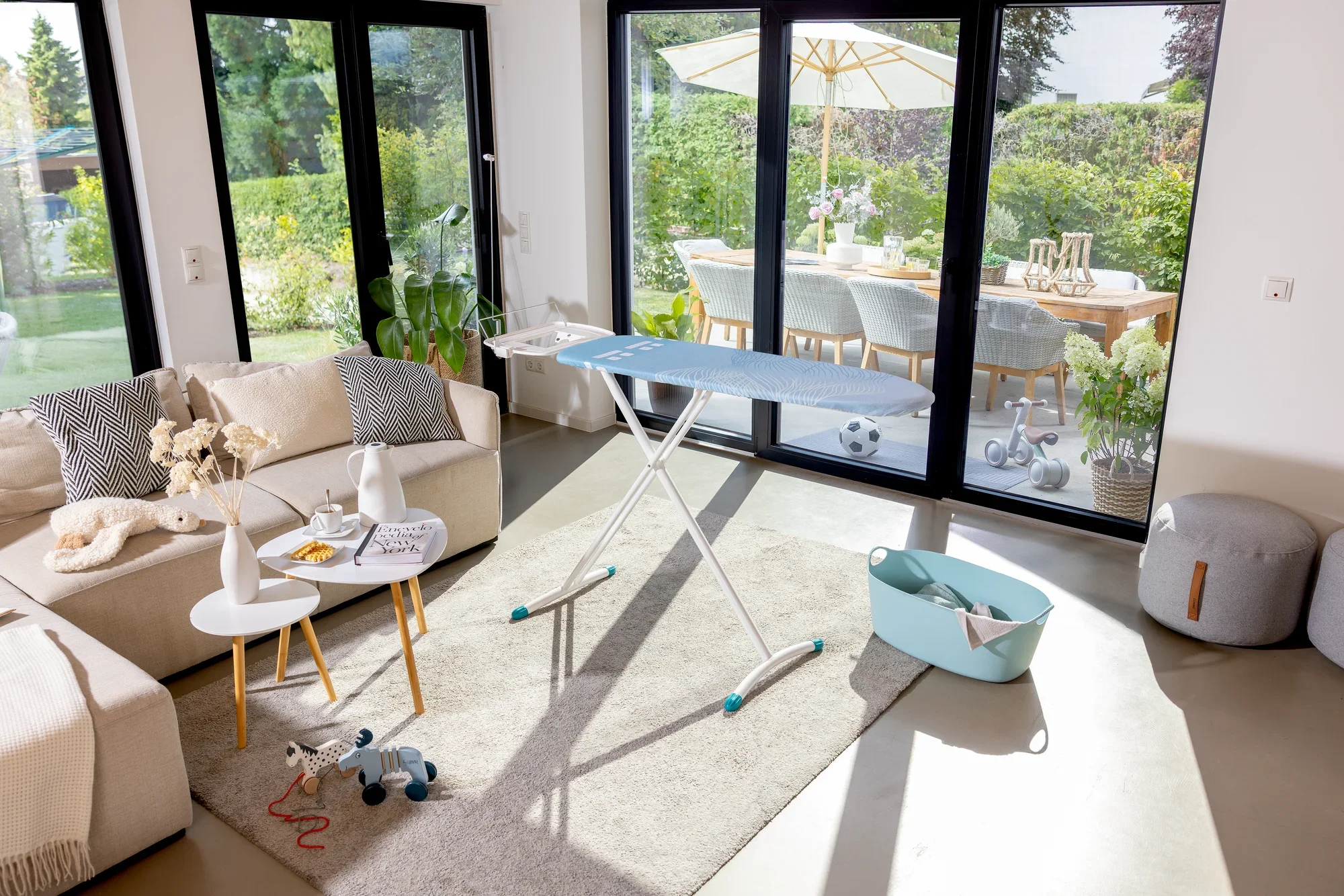 Bügelbrett Leifheit Air Board Express M Solid in einem modernen Wohnzimmer mit großen Fenstern. Es steht auf einem hellen Teppich neben einem Sofa und einem Korb.