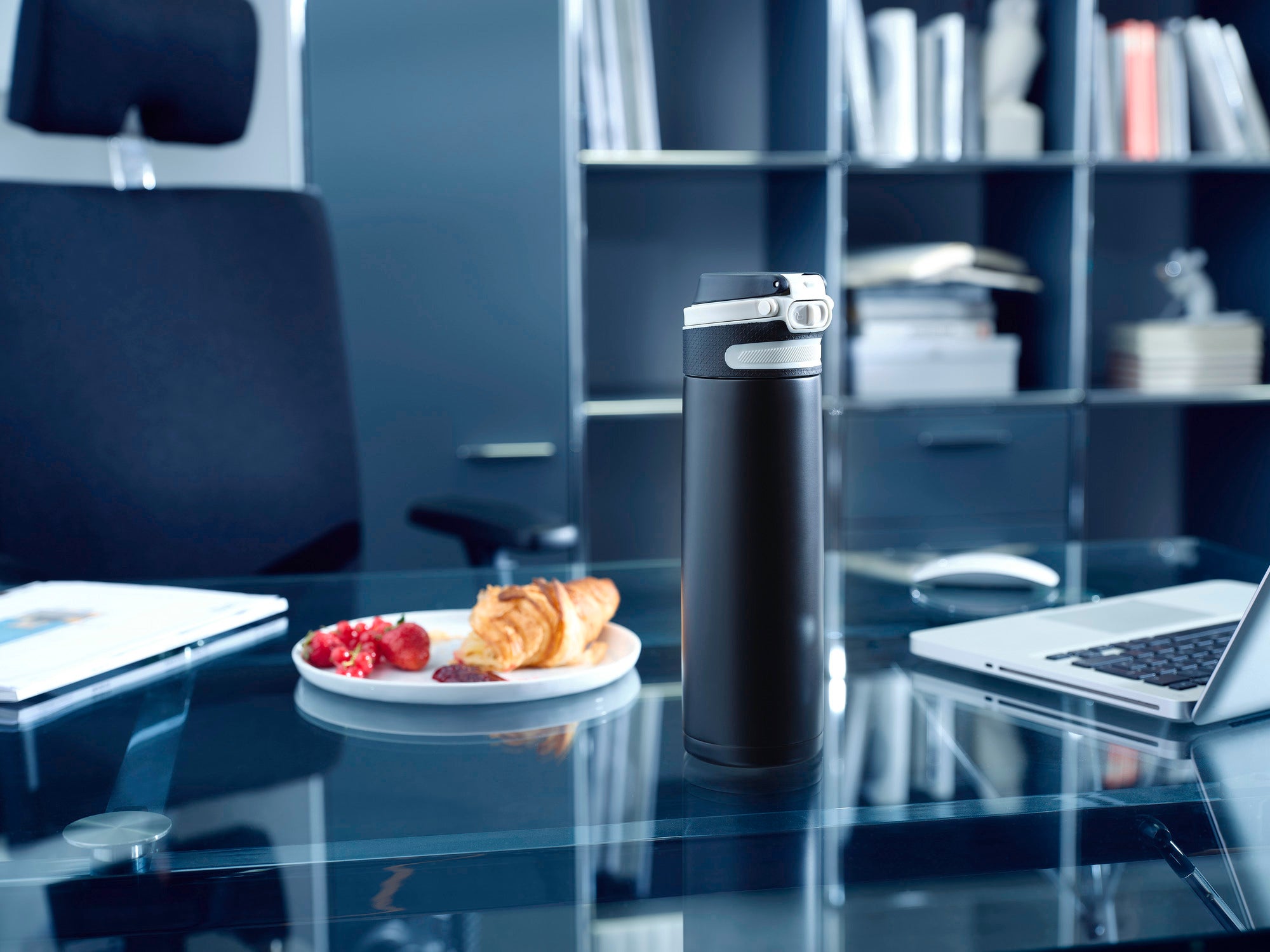 Izolowany kubek Flip 600 ml w kolorze czarnym na nowoczesnym stole biurowym. W tle widoczne są półki z książkami i laptop. Kubek stoi obok talerza z rogalikami i truskawkami.