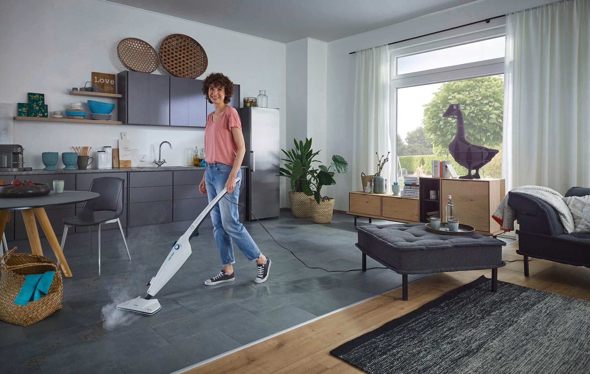 Eine Frau reinigt den Boden einer modernen Küche mit dem Dampfreiniger CleanTenso Power. Der Raum ist hell und stilvoll eingerichtet, mit Pflanzen und Dekorationen.