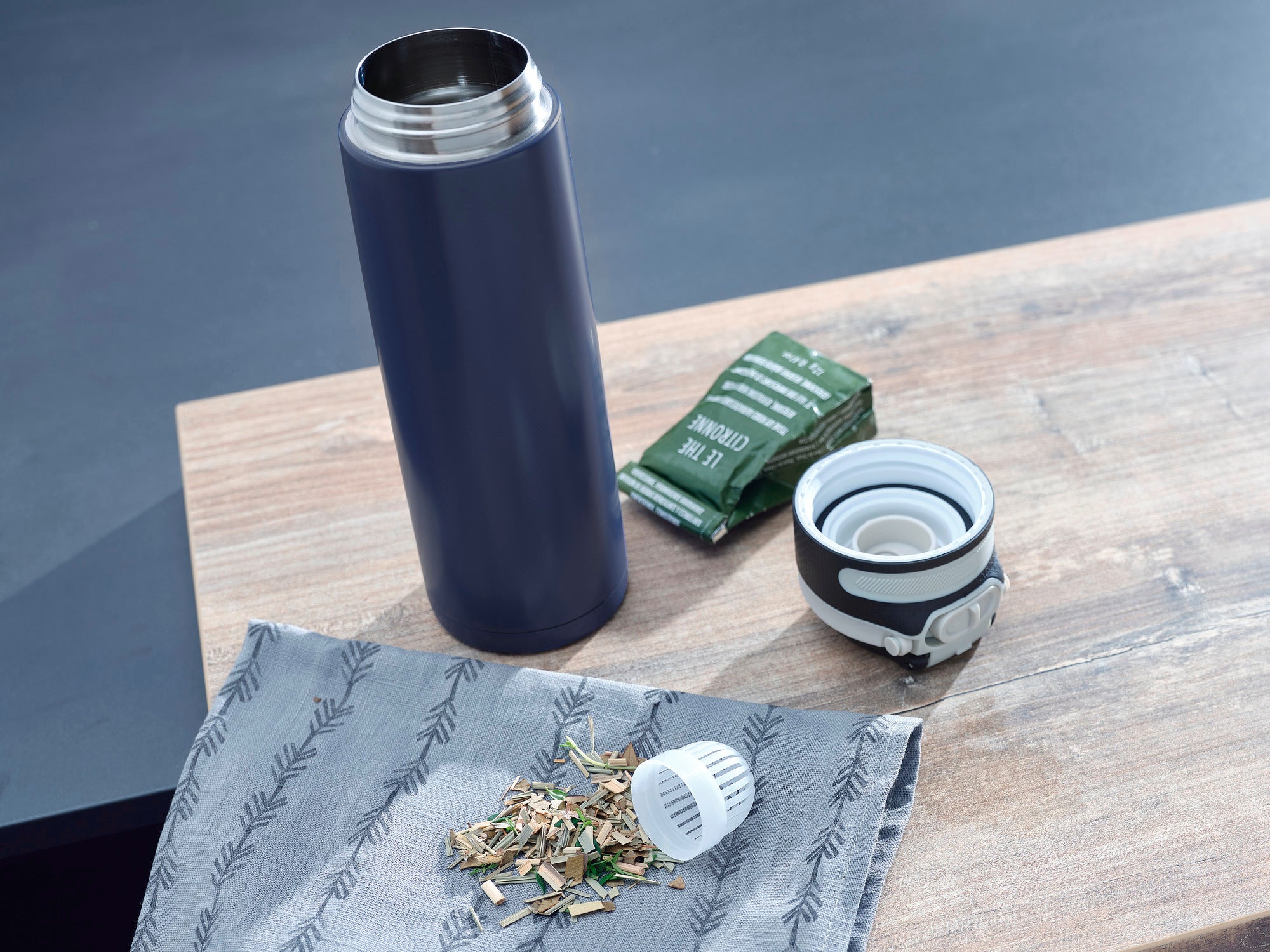 Isolierbecher Flip 600ml in Dunkelblau auf einem Tisch. Der Becher ist geöffnet, daneben liegen ein Teebeutel, ein Deckel und lose Teeblätter auf einem Tuch.
