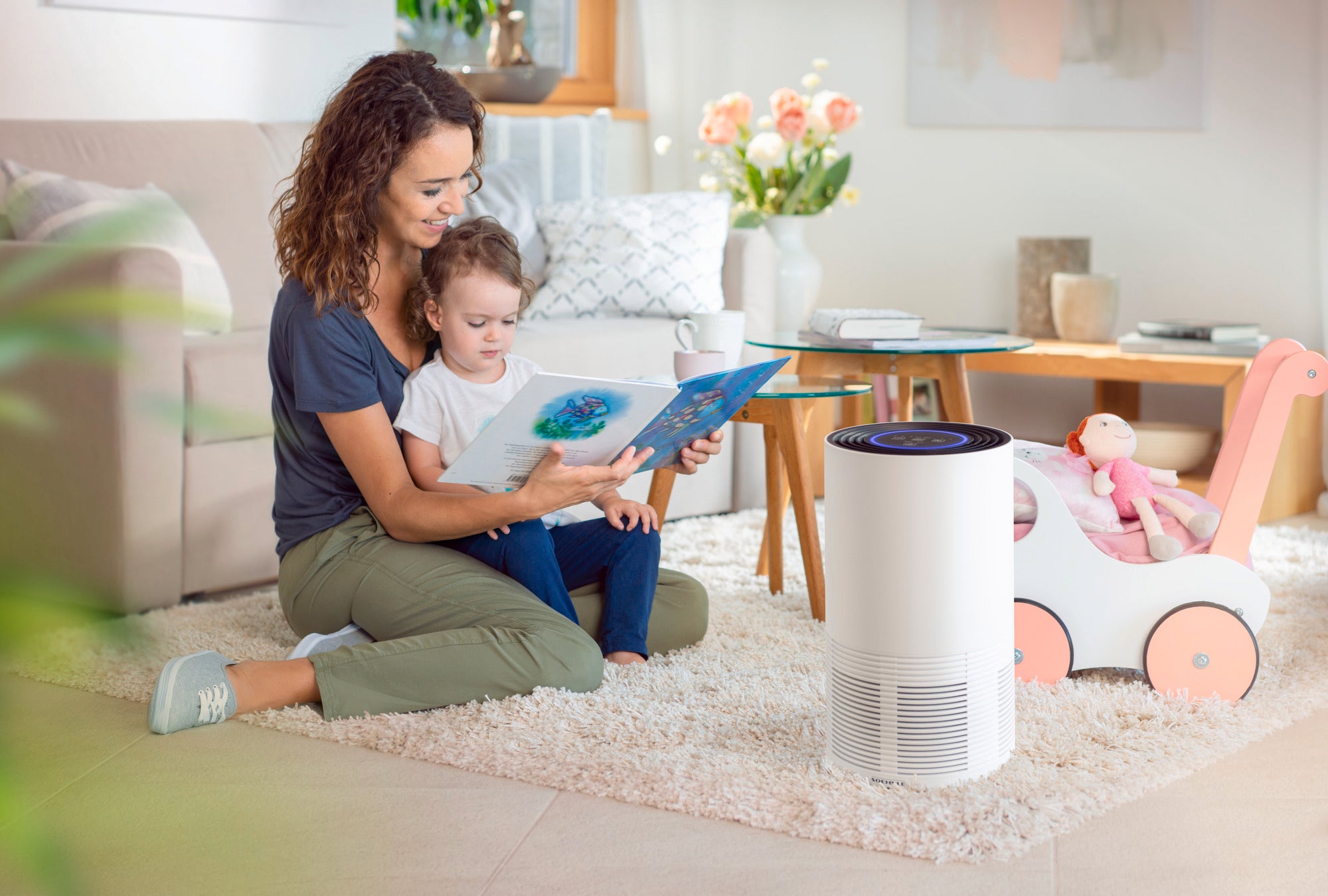 Een moeder leest een boek voor aan haar kind in de woonkamer. Op de voorgrond staat de Airfresh Clean 400 luchtreiniger, die zorgt voor schone en frisse lucht.