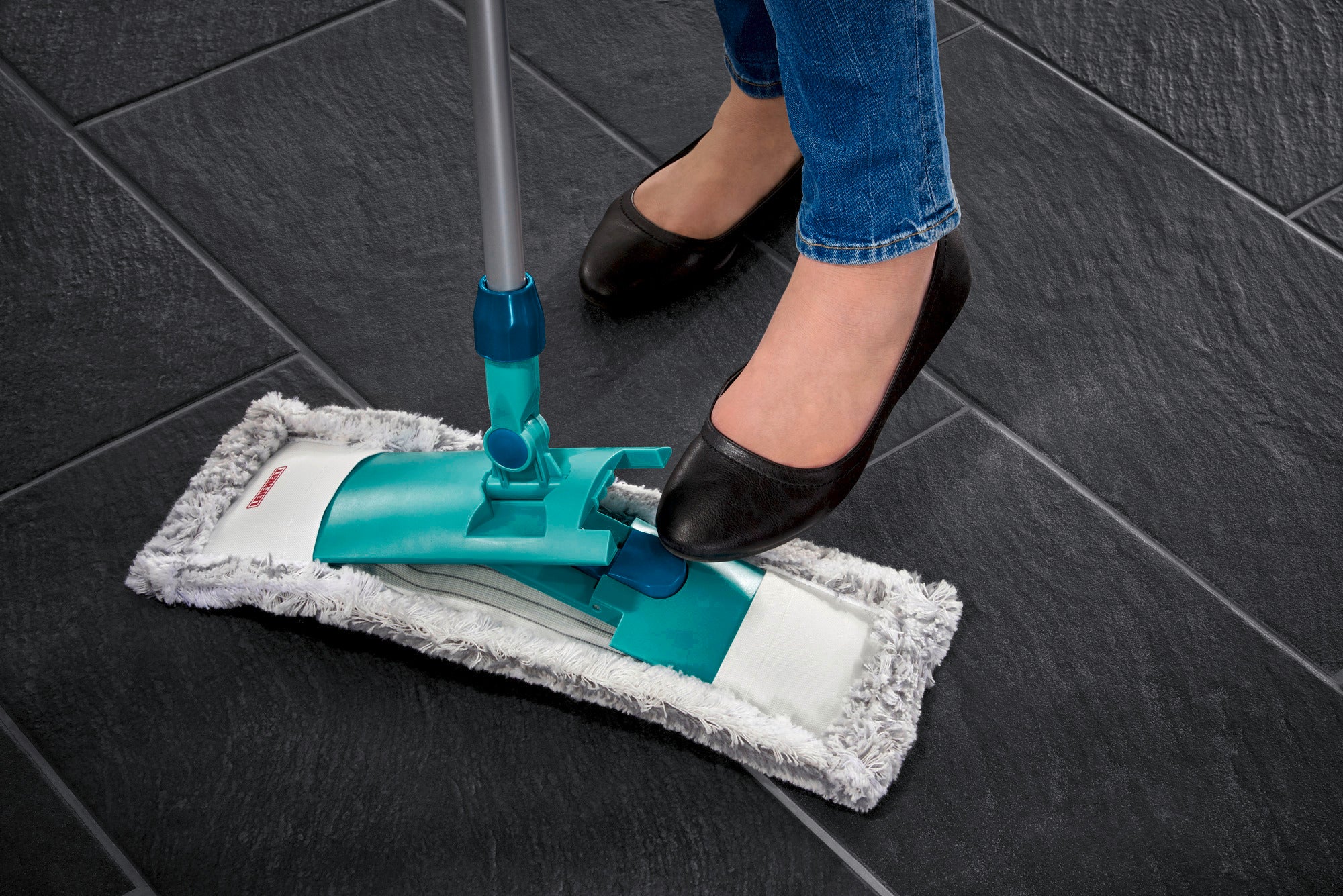 Een persoon gebruikt de Classic vloerwisser op donkere tegels. De mop heeft een groene steel en een grijze mopdoek die wordt bediend met een voetpedaal.