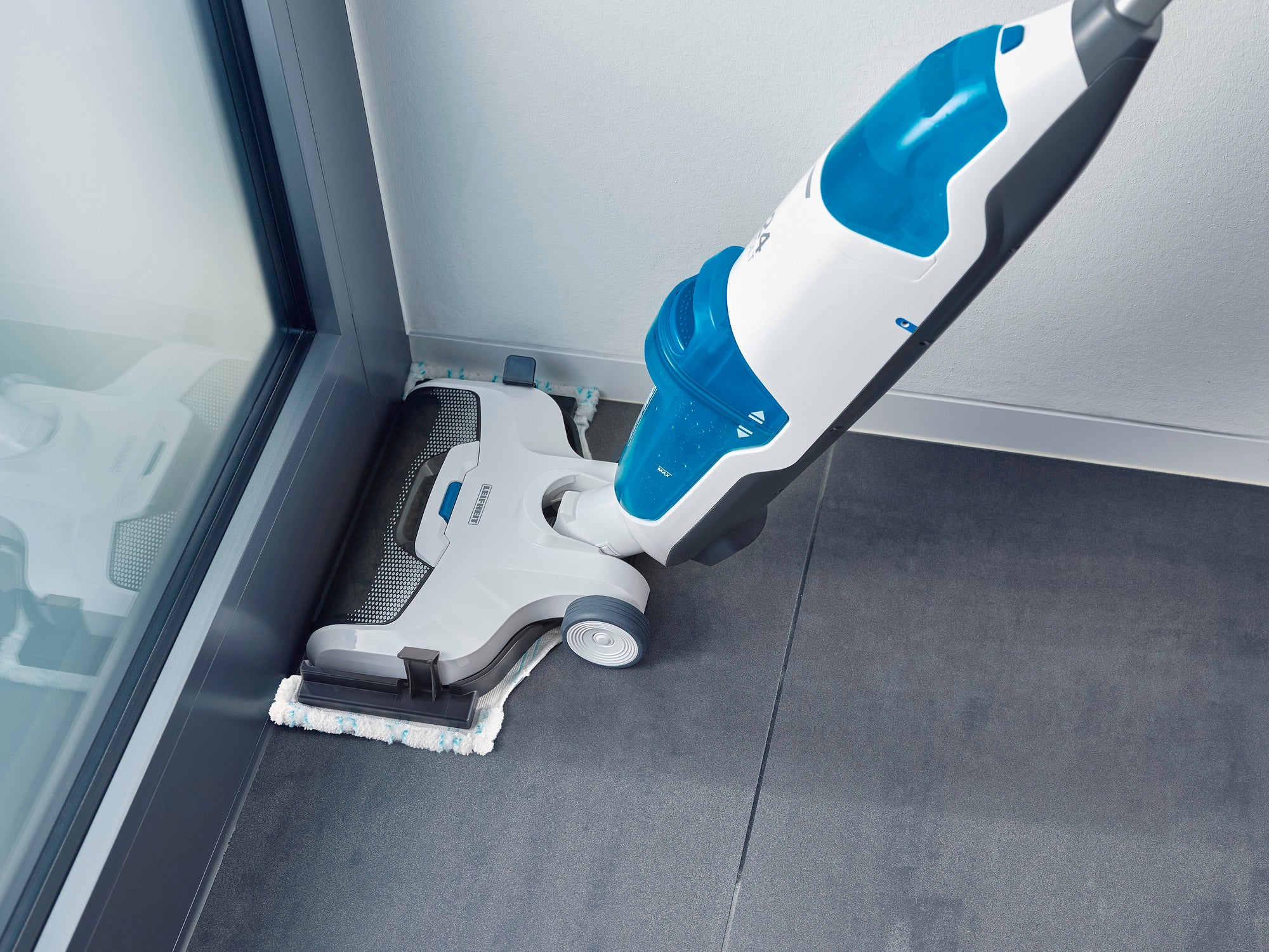 Aspirateur Regulus Aqua PowerVac Pro en gros plan, nettoyant un sol gris près d'une fenêtre. L'appareil est blanc et bleu, conçu pour atteindre les coins difficiles.