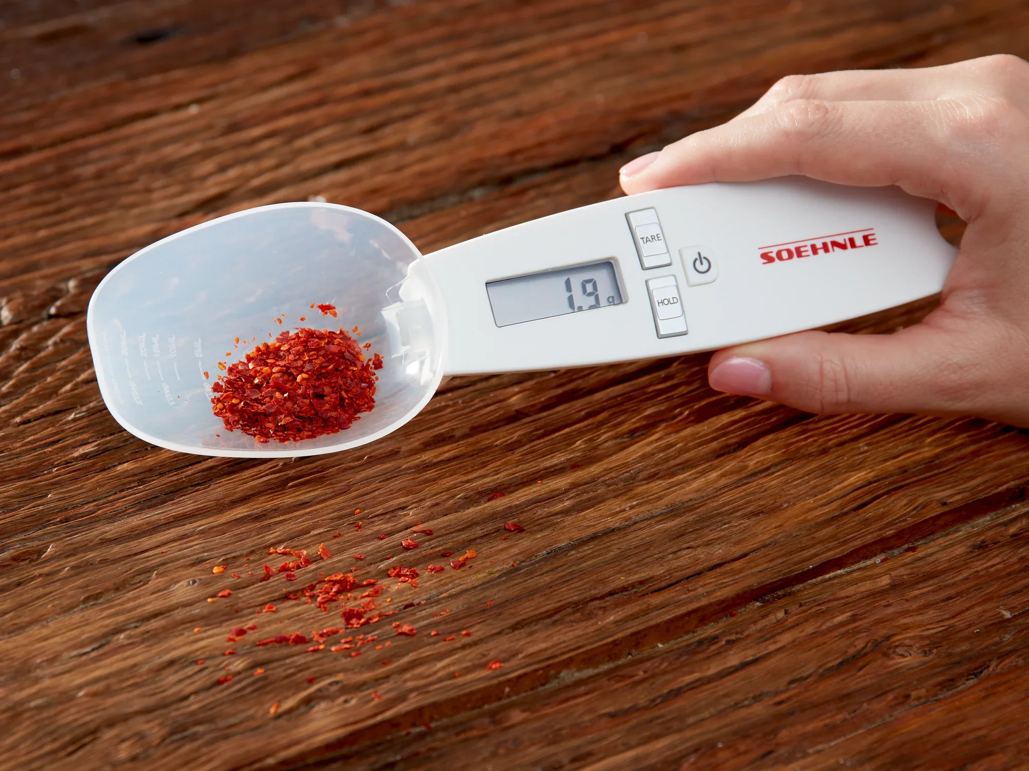 Ręka trzyma cyfrową wagę kuchenną Cooking Star firmy Soehnle. Waga wyświetla 1,9 grama i waży czerwone przyprawy na drewnianej podstawie. Idealna do precyzyjnych pomiarów.