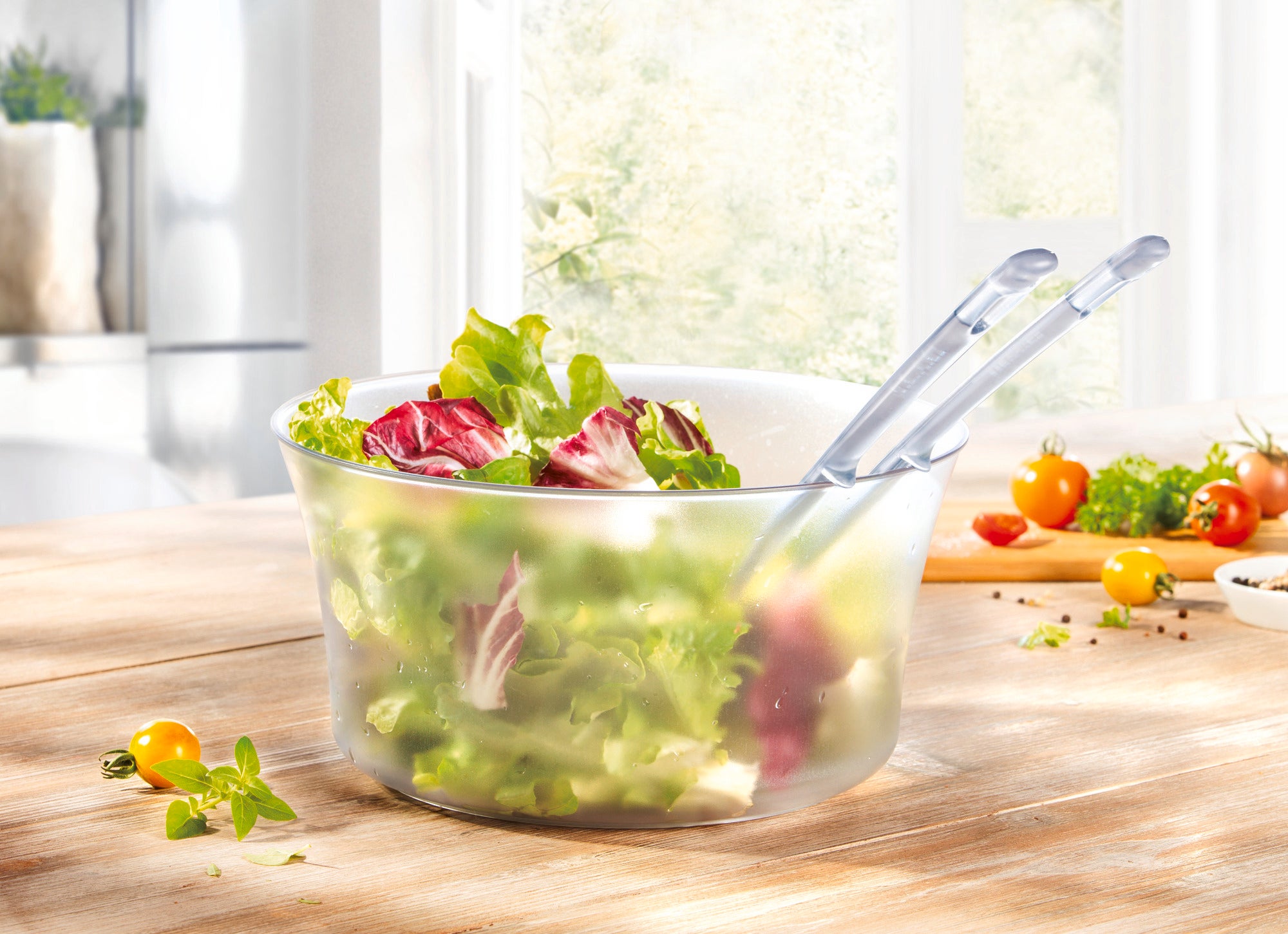 Salatbesteck in einer transparenten Schüssel mit frischem, grünem Salat und roten Radicchio-Blättern. Im Hintergrund sind Tomaten und Kräuter auf einem Holztisch zu sehen.