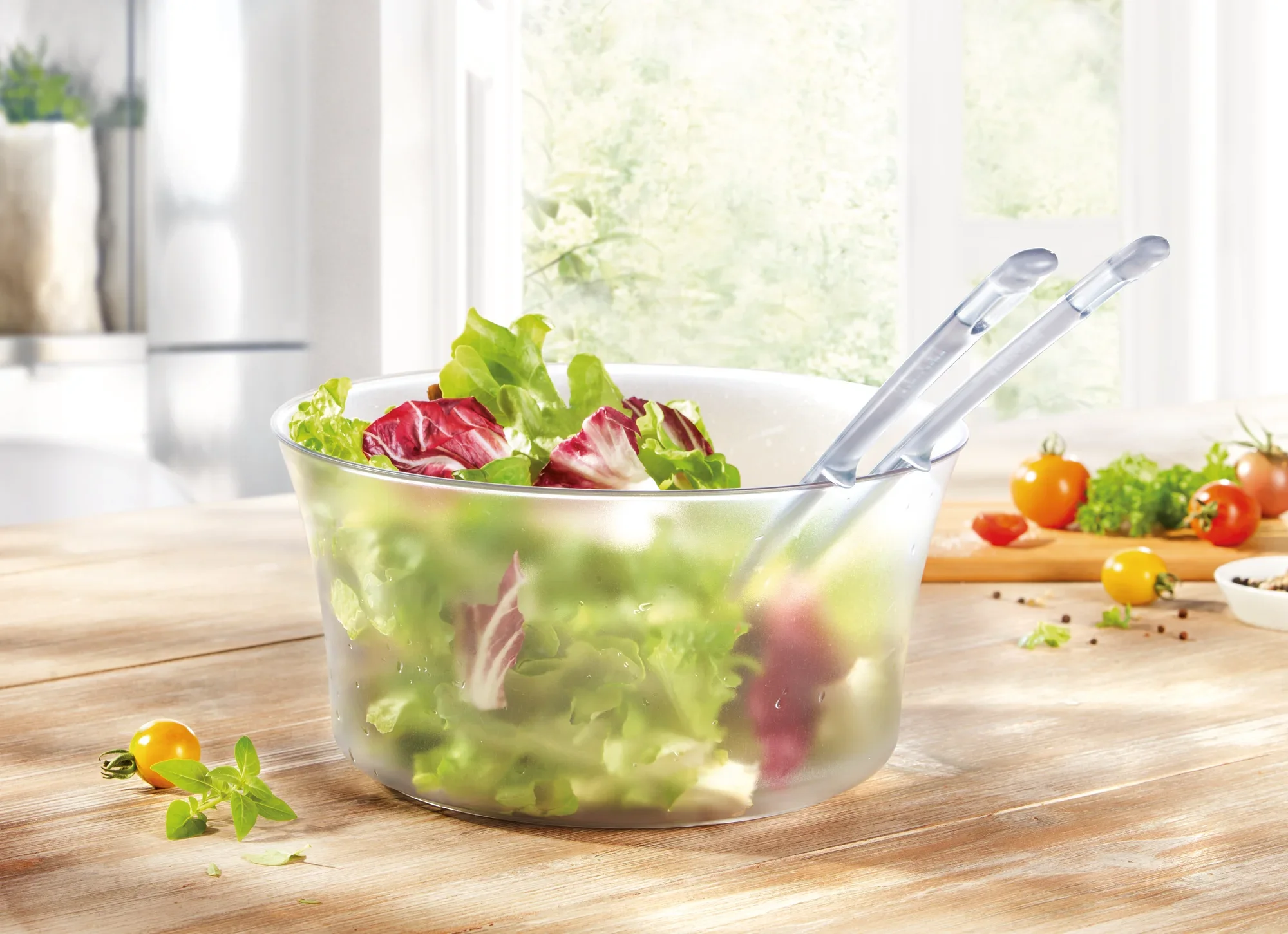 Cubiertos para ensalada en un cuenco transparente con lechuga fresca y hojas de achicoria roja. Al fondo se ven tomates y hierbas sobre una mesa de madera.
