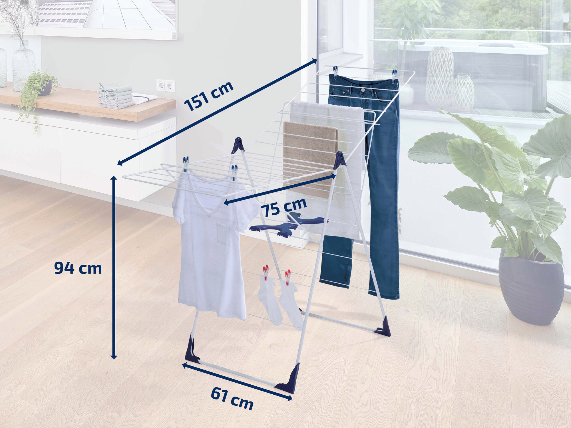 Een vrijstaande wasdroger van Leifheit in een moderne kamer. De droger heeft een hoogte van 94 cm, een breedte van 61 cm en een lengte van 151 cm. Hij is geladen met kleding en staat naast een plant.