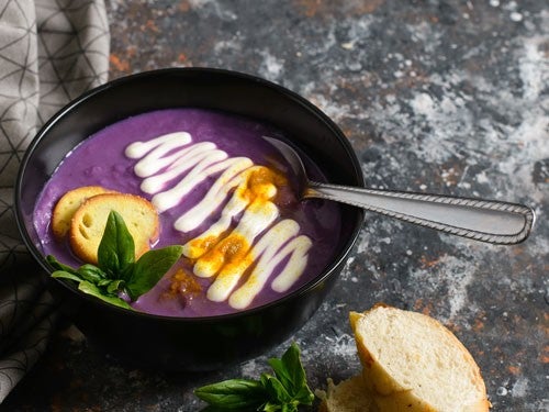 Eine schwarze Schuessel mit violetter Rotkohl-Suppe, verziert mit Sahne, Brotchips und Basilikum, daneben ein Stueck Baguette.