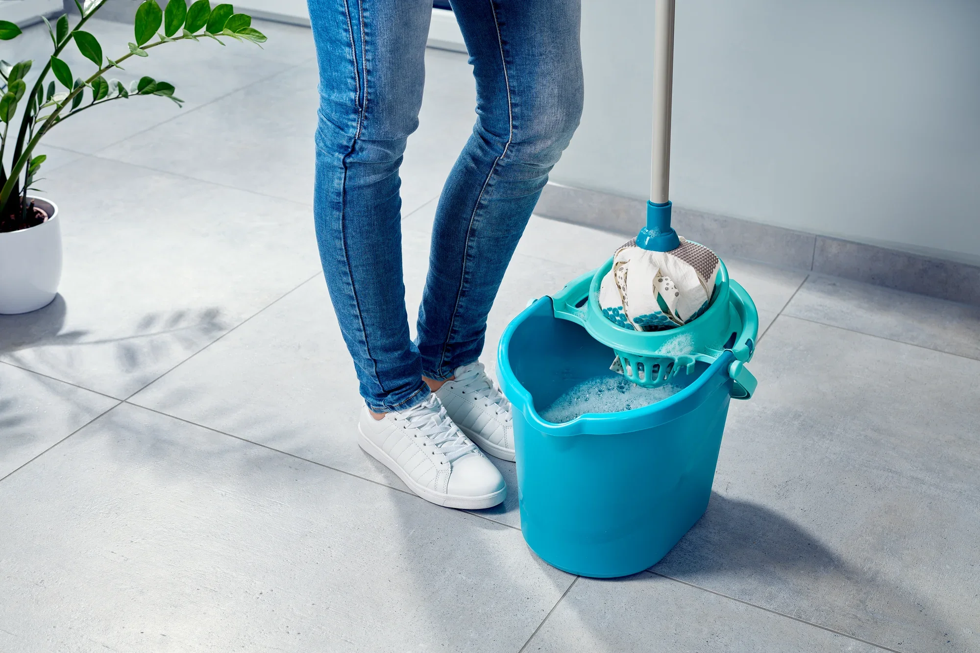 A person is standing on a tiled floor next to a bucket containing a Power Mop 3in1. The mop head is immersed in the bucket, ready for cleaning. A plant can be seen in the background.