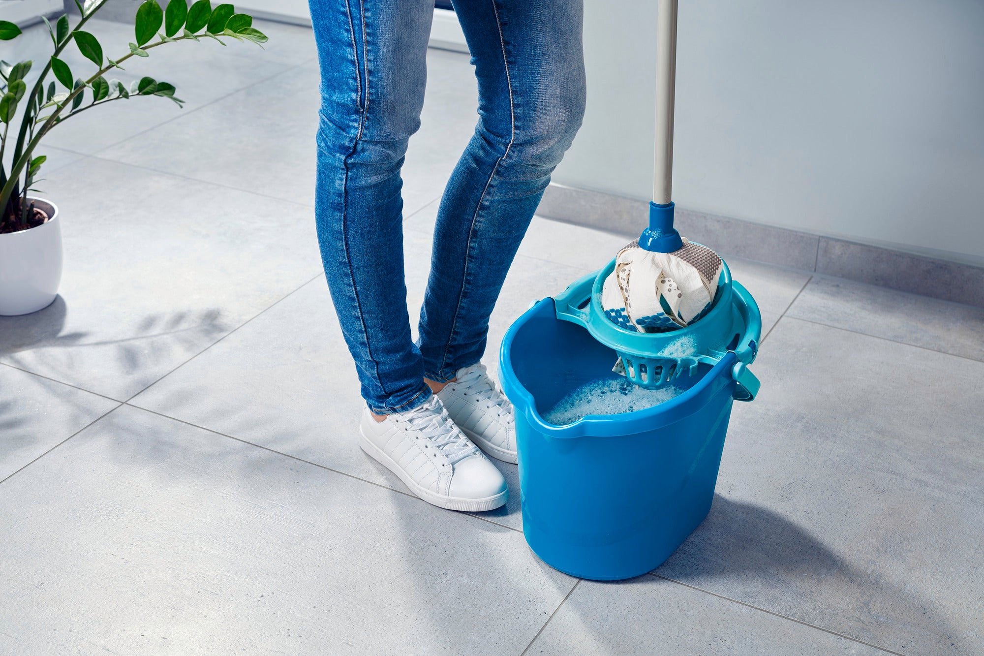 Una persona está de pie sobre un suelo de baldosas junto a un cubo con una Power Mop 3in1. El cabezal de la mopa está sumergido en el cubo, listo para limpiar. Al fondo se ve una planta.