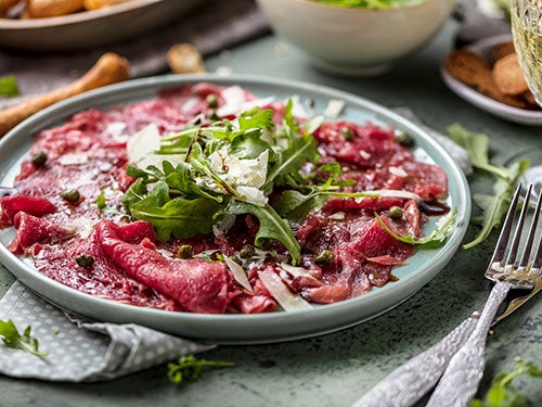 Ein Teller mit Carpaccio aus rohem Rindfleisch, garniert mit Rucola, Kapern und Parmesanspaenen, daneben Besteck auf grauem Untergrund.