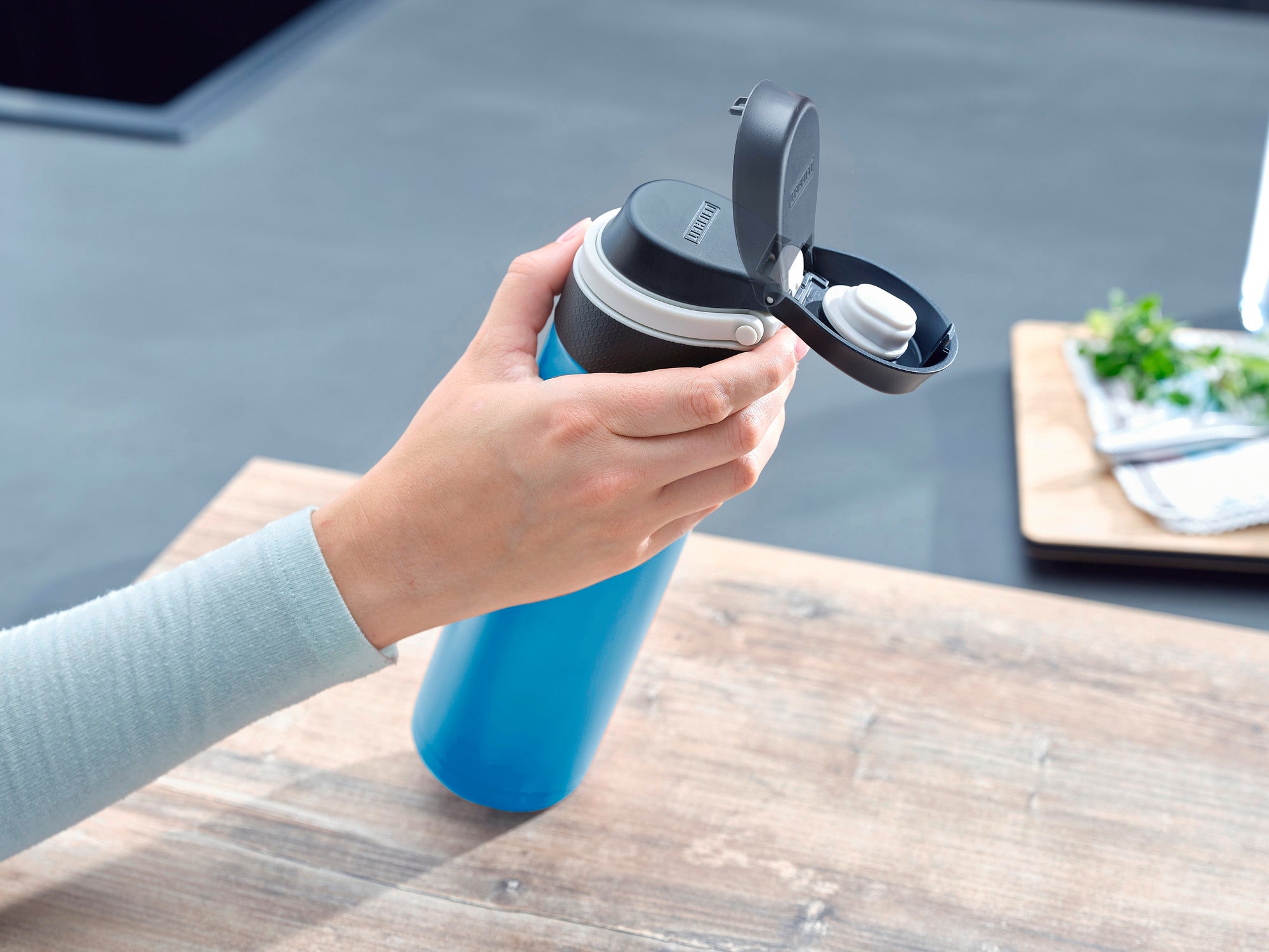 Isolierbecher Flip 600ml in Hellblau wird auf einem Tisch verwendet. Eine Hand öffnet den Deckel des Bechers, ideal für unterwegs, um Getränke warm oder kalt zu halten.
