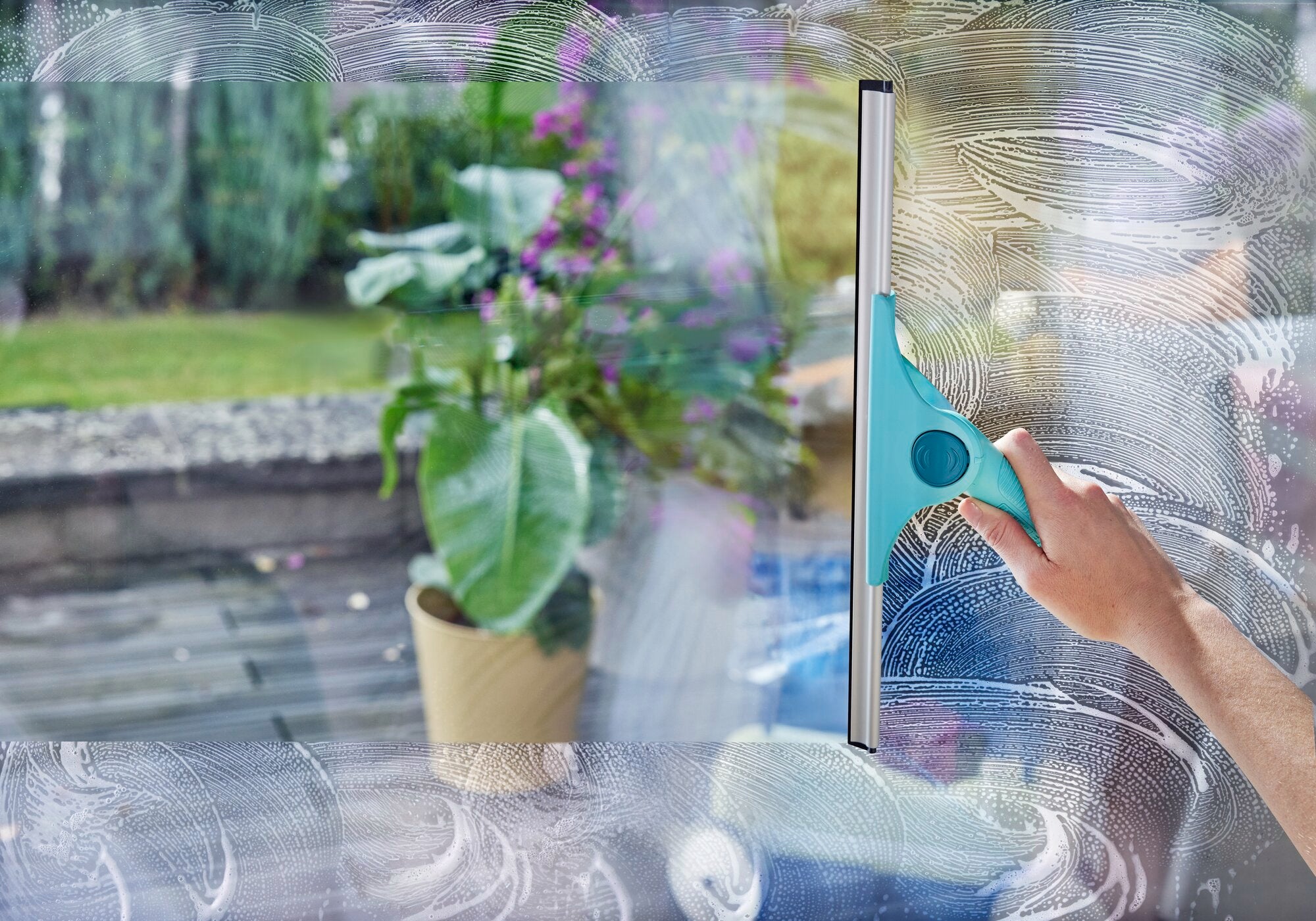 Ein Fensterreiniger namens "Window Slider 2in1 XL ProReach" wird verwendet, um ein großes Fenster zu reinigen. Im Hintergrund ist ein Garten mit Pflanzen sichtbar.