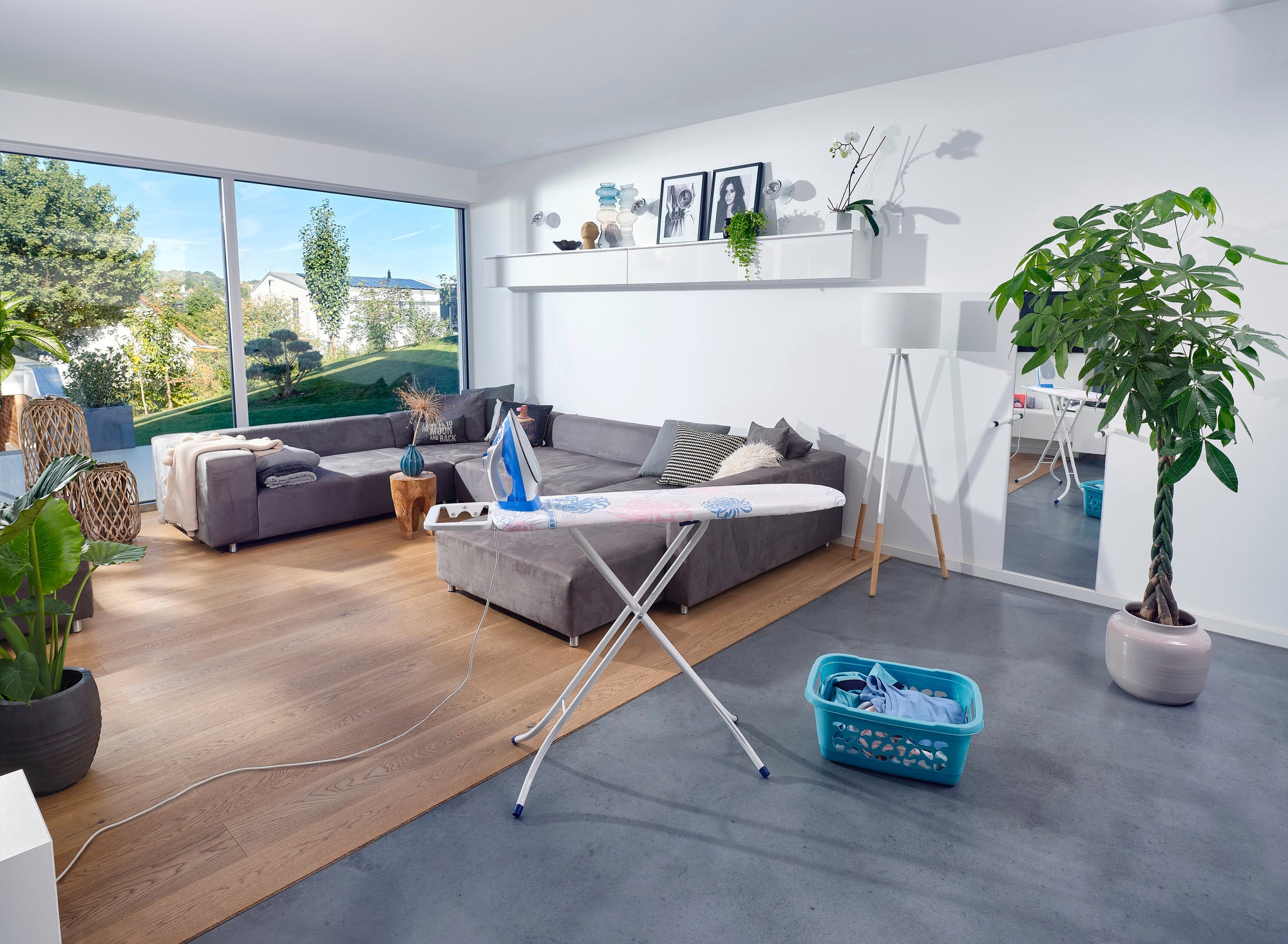 Bügelbrett Classic S Basic in einem modernen Wohnzimmer mit großen Fenstern, grauem Sofa und Pflanzen. Das Bügelbrett steht bereit, um Kleidung zu glätten.