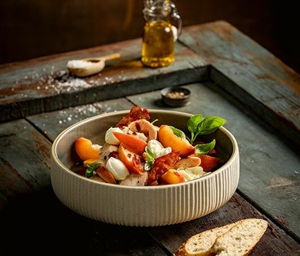 Ein rustikaler Holztisch mit einem Salat aus Pfirsichen, Bueffelmozzarella, Parmaschinken, Haehnchen und Basilikum, daneben frisches Brot und Olivenoel.